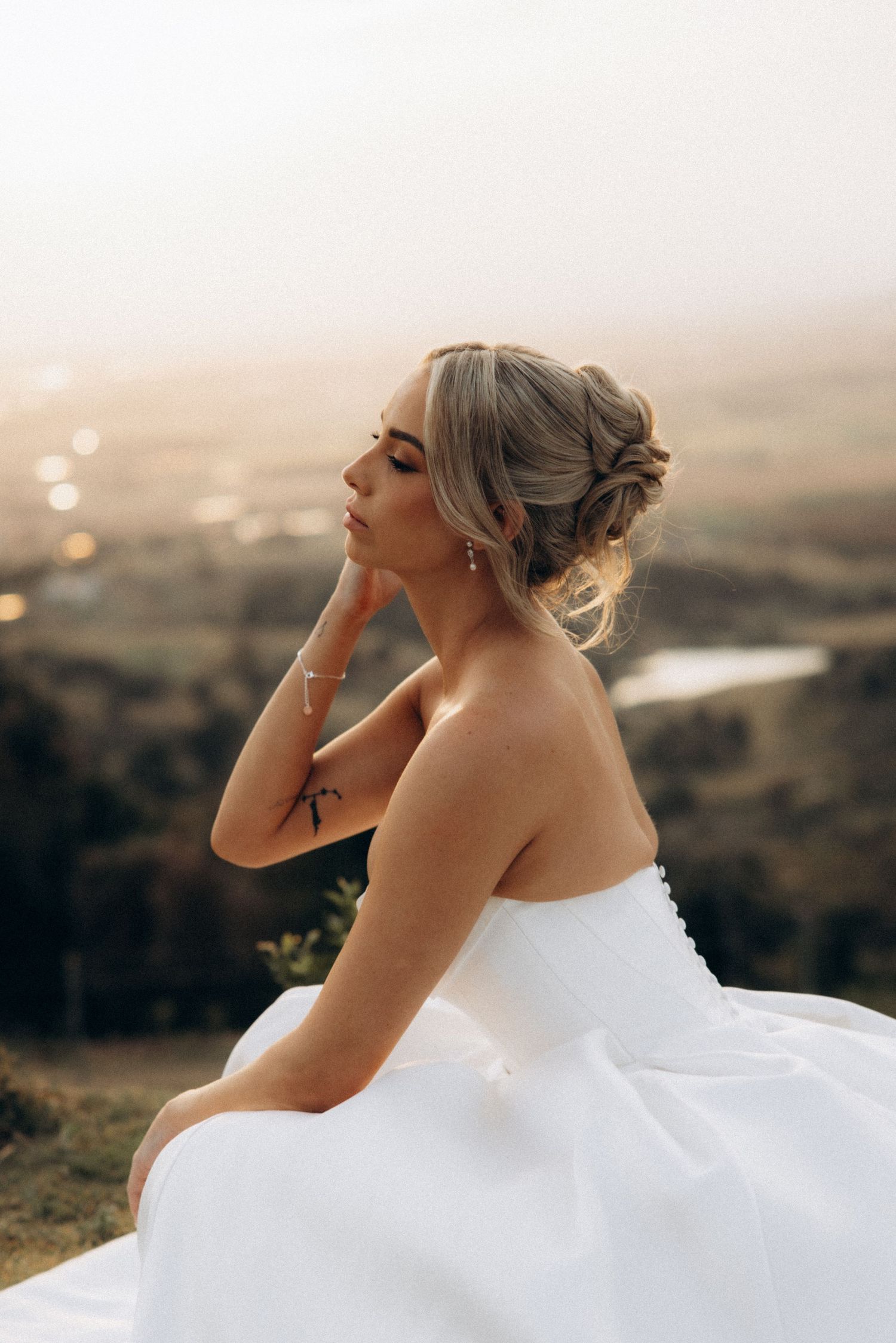 Bride in a flowing white wedding dress poses dramatically against a scenic sunset landscape.