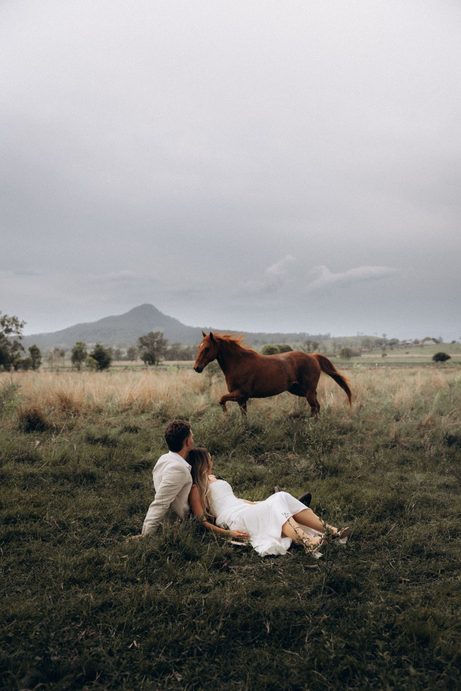 A couple relaxes in a field while horses graze in the background.