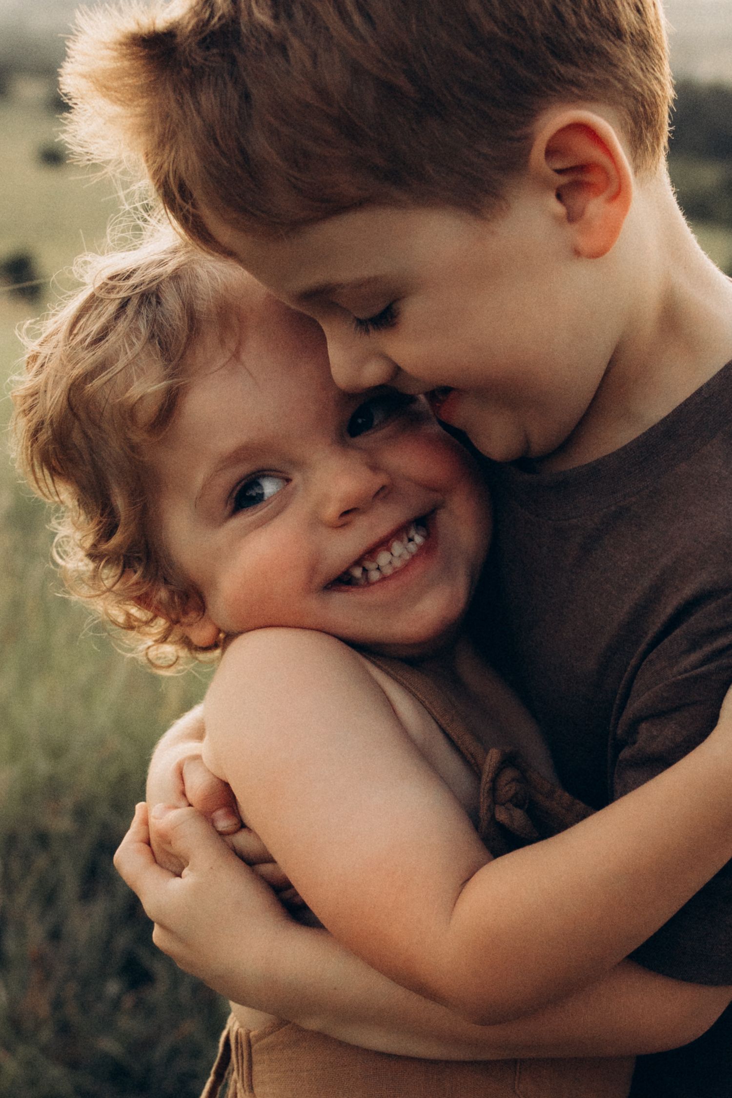 Two children share a tender embrace while playing outdoors in a grassy field at sunset.