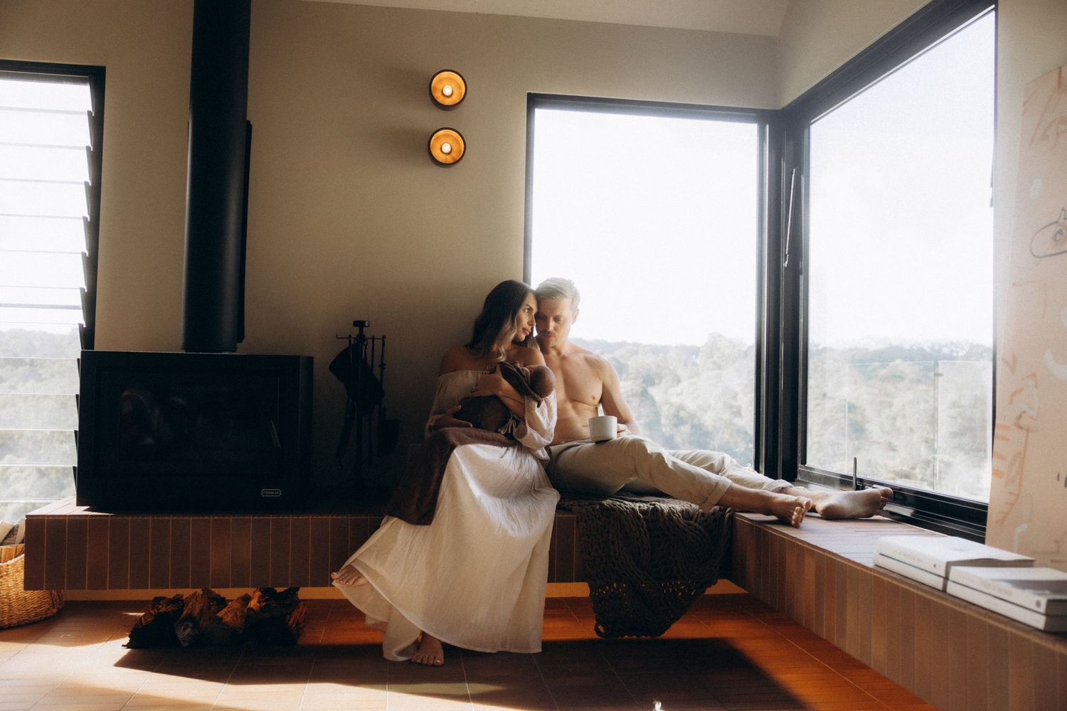 Parents and newborn relaxing by window in sunlit modern interior.