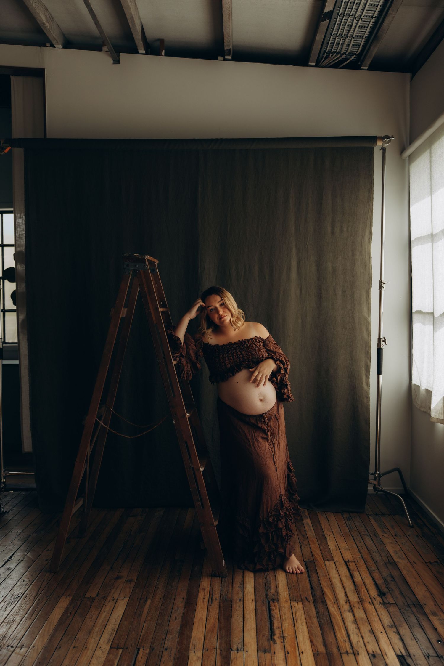 Pregnant woman in brown outfit poses by wooden ladder against dark backdrop in a photography studio.