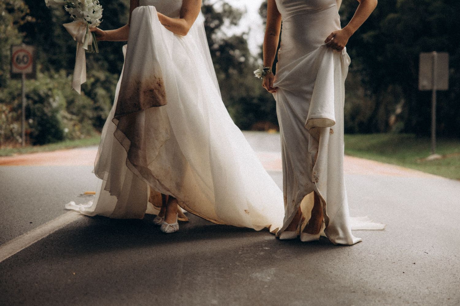 Bride and bridesmaid lifting white wedding gowns while walking on road, showing muddy hems and heels.