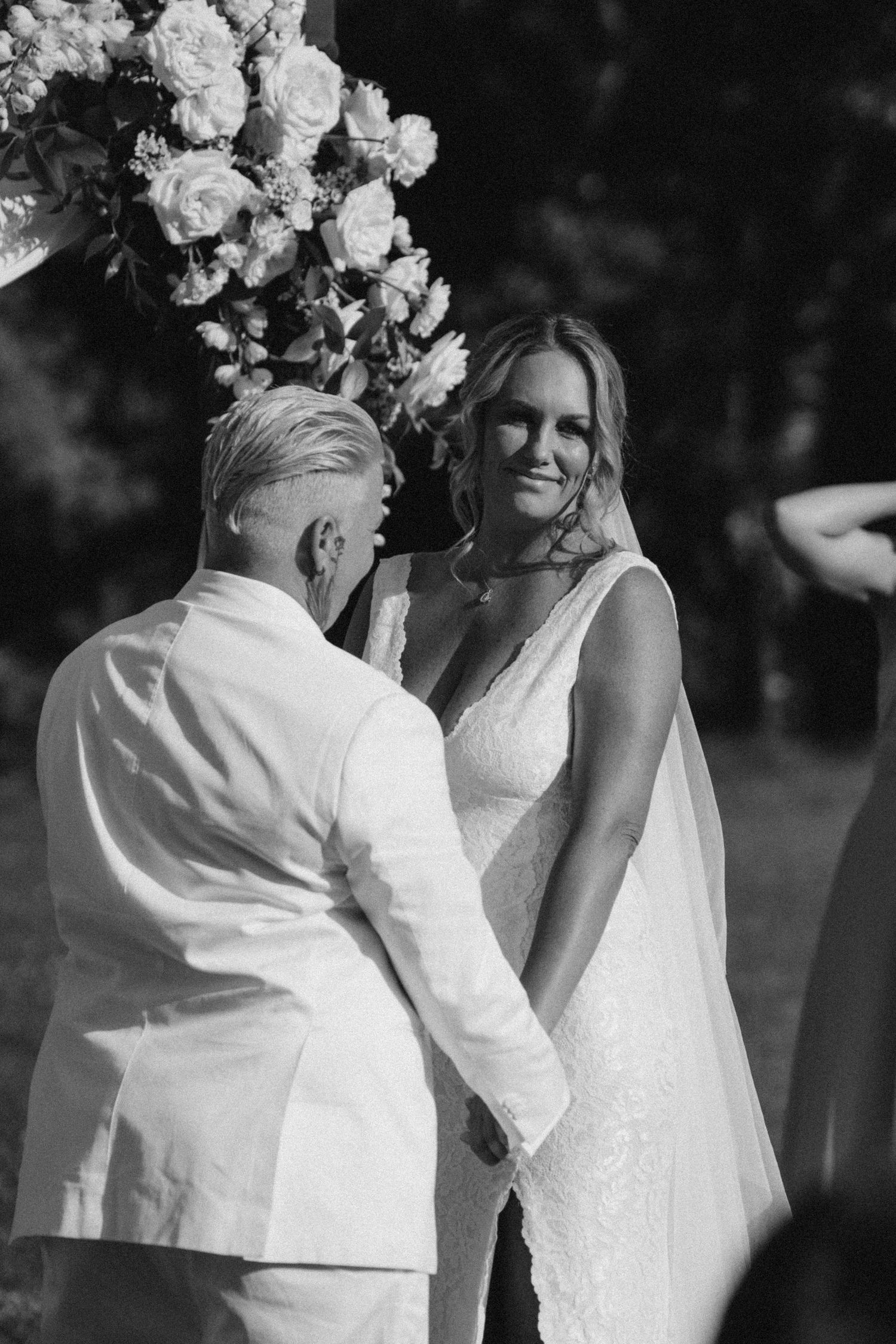 Black and white wedding photo of two brides holding hands at outdoor ceremony beneath floral arch.