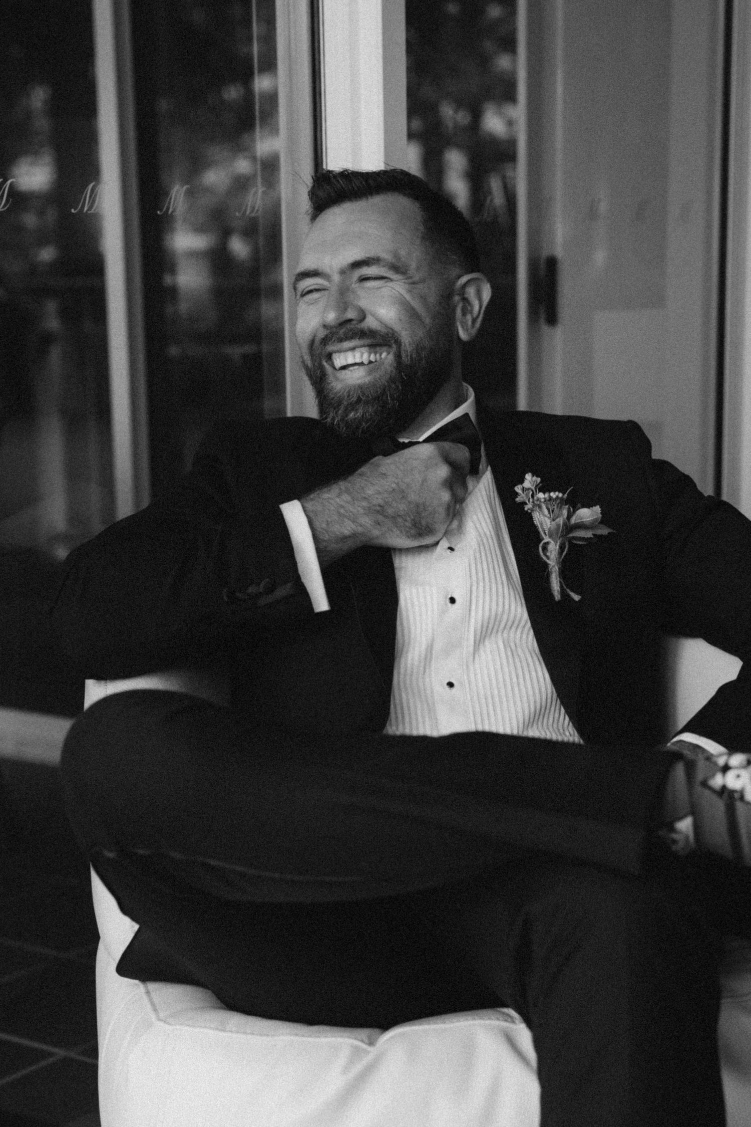 Laughing groom in black tuxedo with boutonniere sits relaxed in chair, candid black and white wedding portrait.