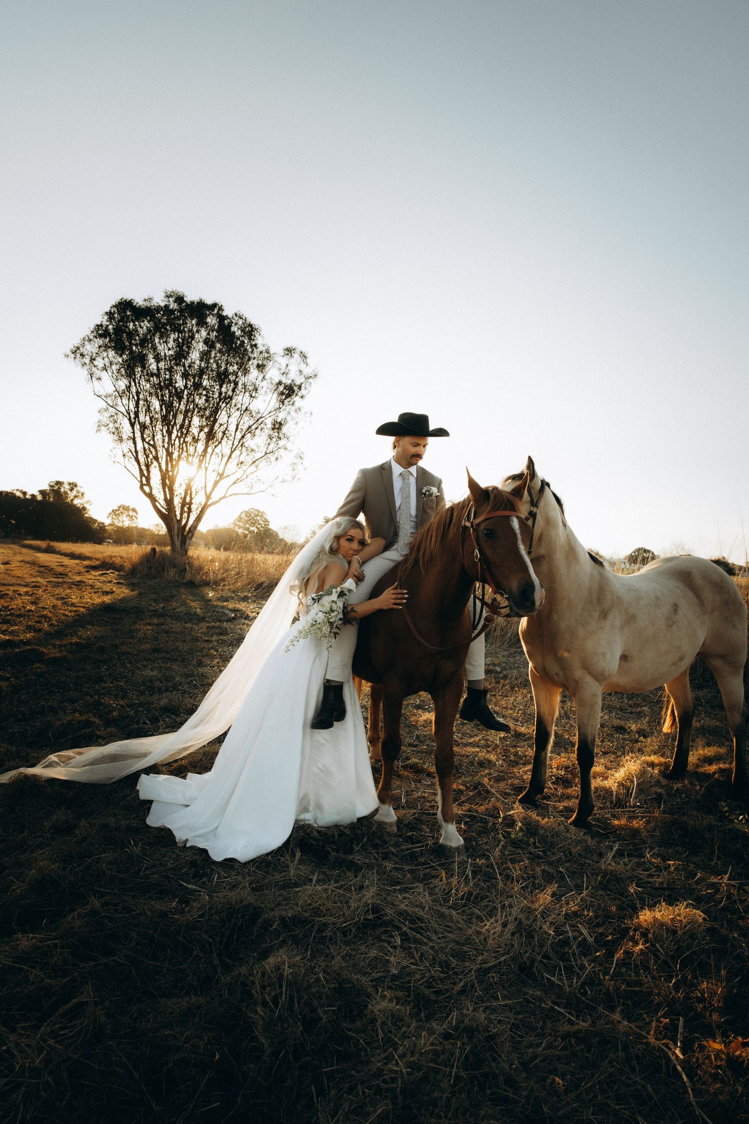 Bride and groom with horses at sunset on a rustic farm, groom wearing cowboy hat, bride in flowing white gown.