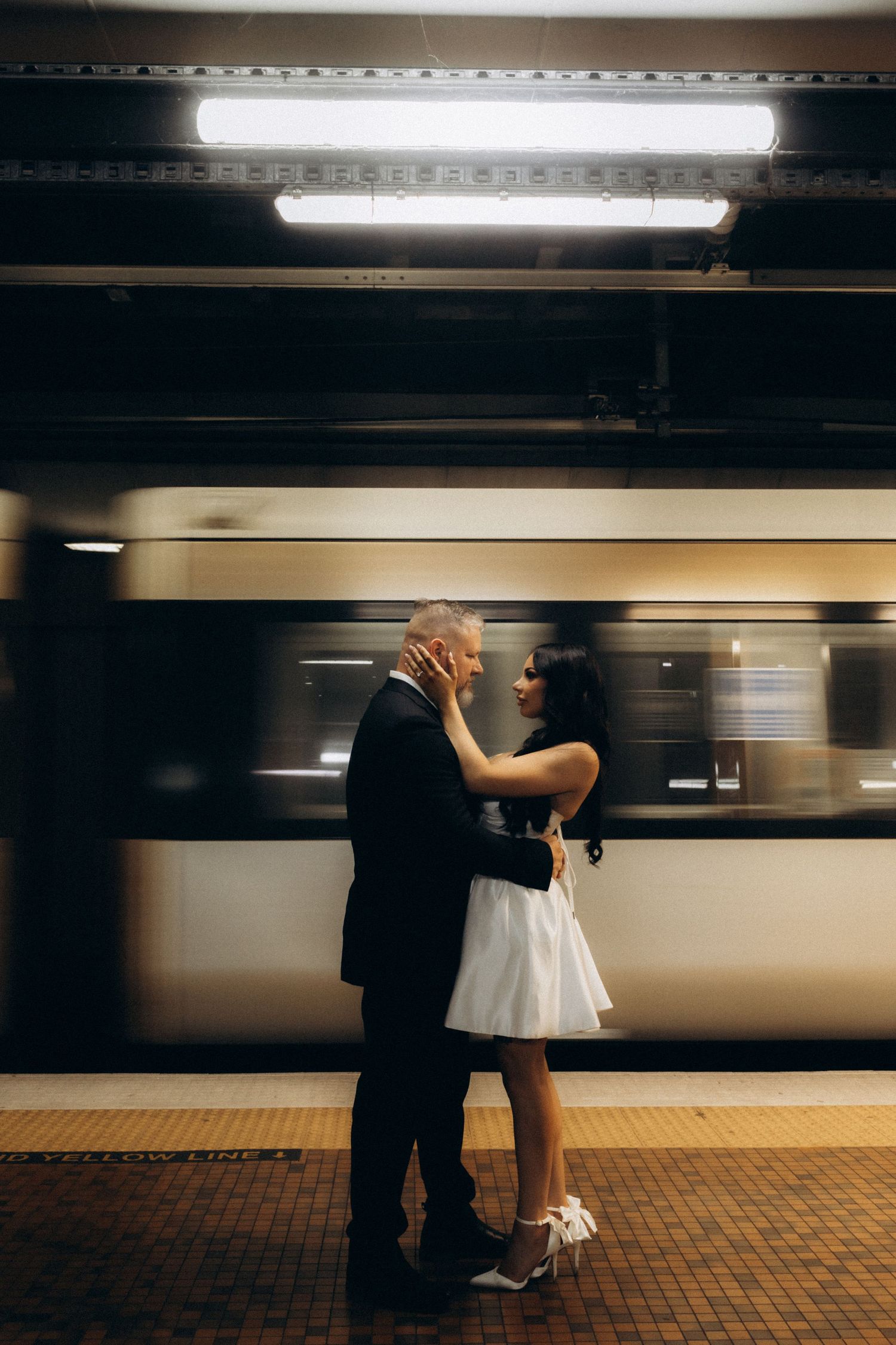 Couple embracing on subway platform in wedding attire with a blurred train passing behind them.