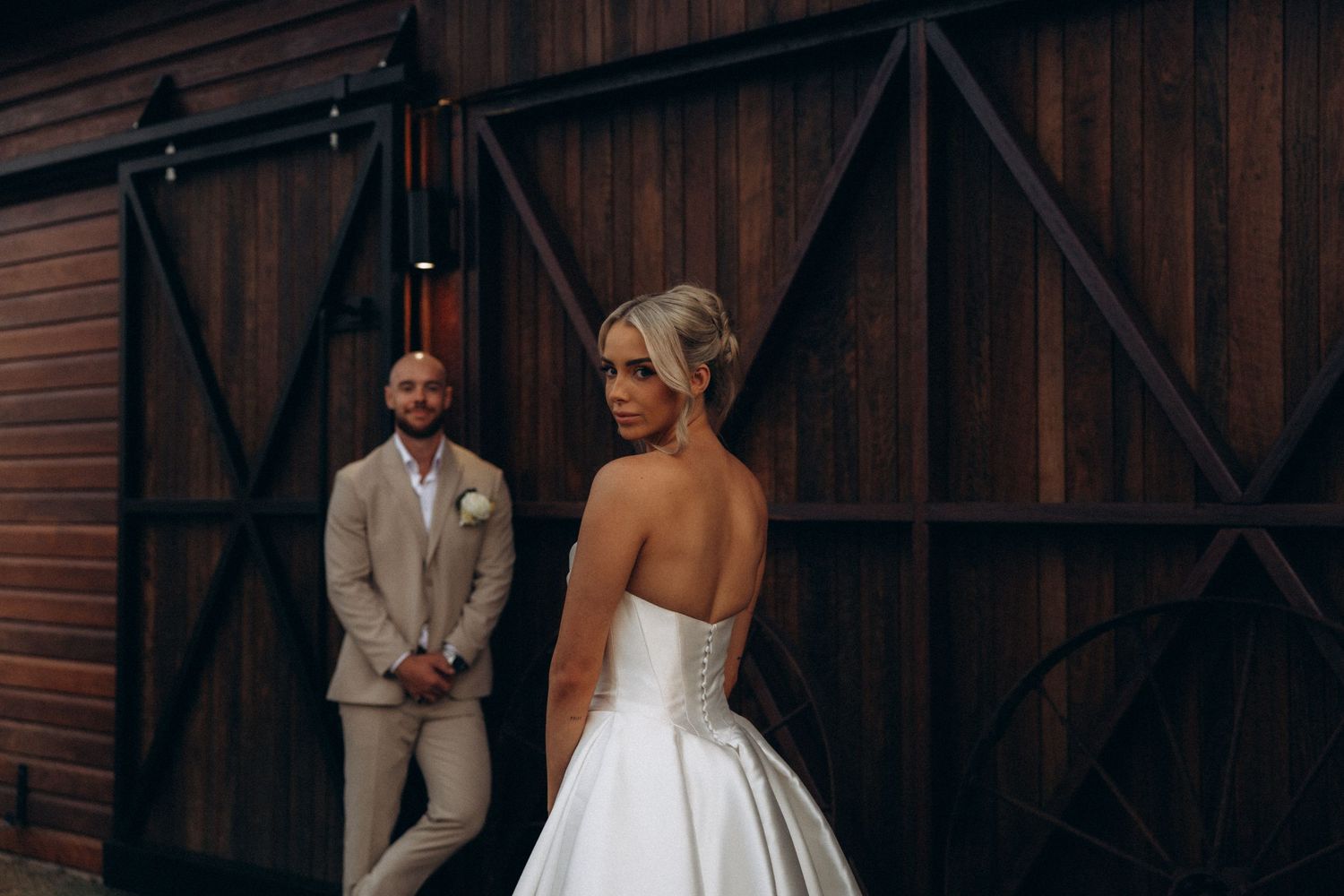 Couple in wedding attire shares candid moments by a rustic barn wall.