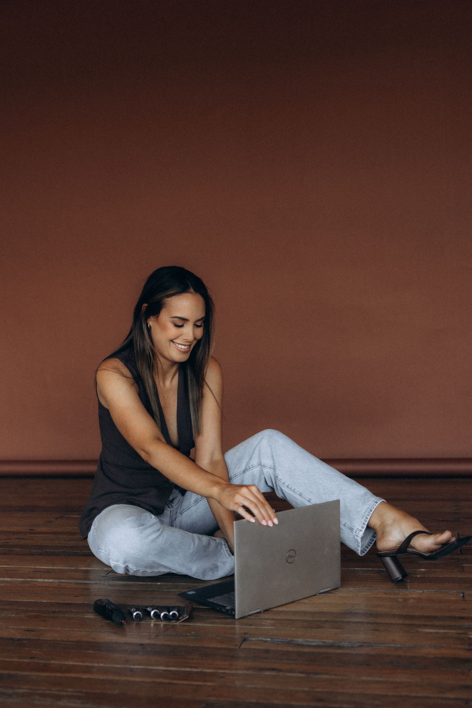 Person in casual attire working on laptop while relaxing on wooden floor with brown background.
