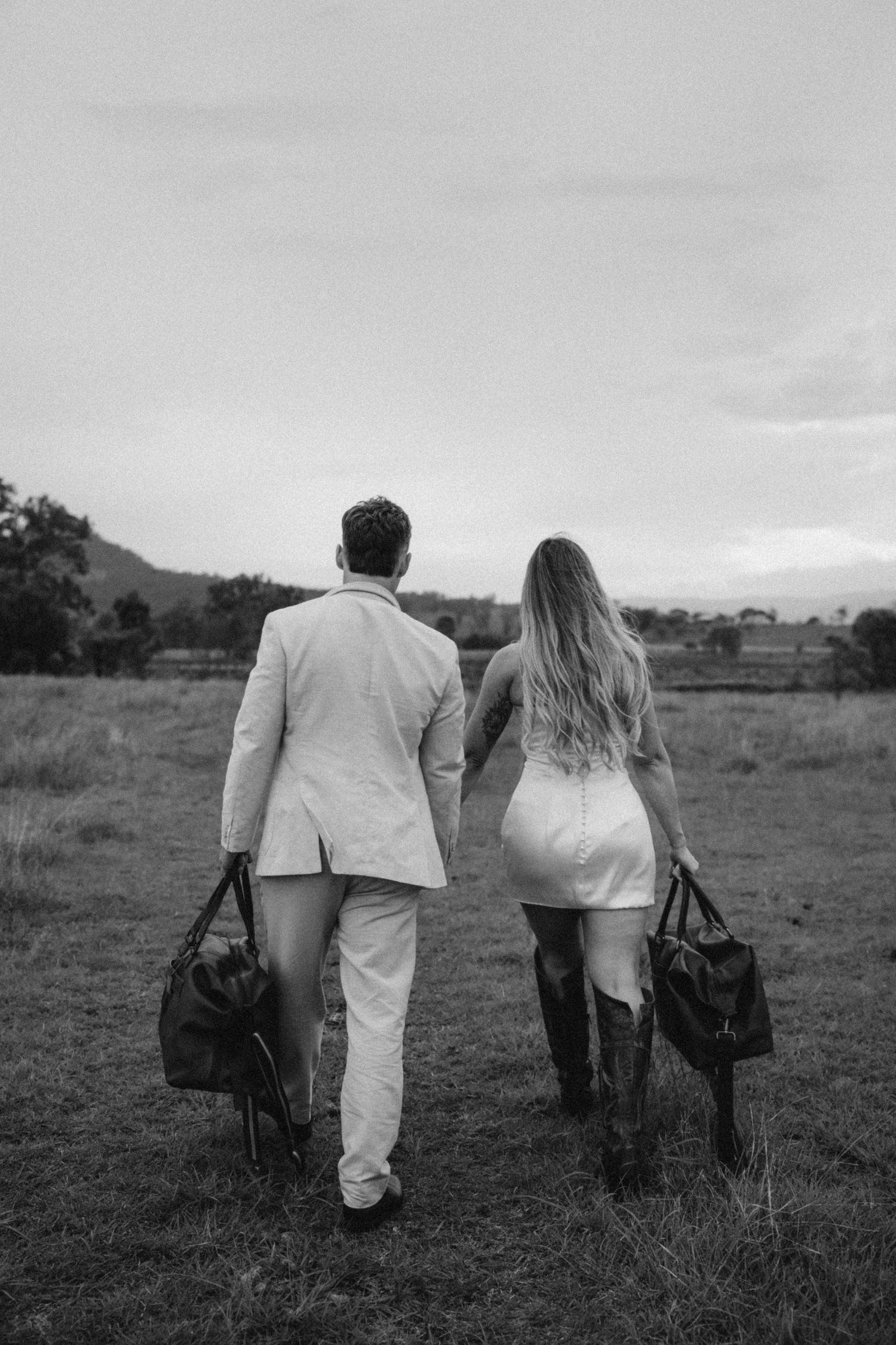 A couple walking away from camera carrying bags across a field.
