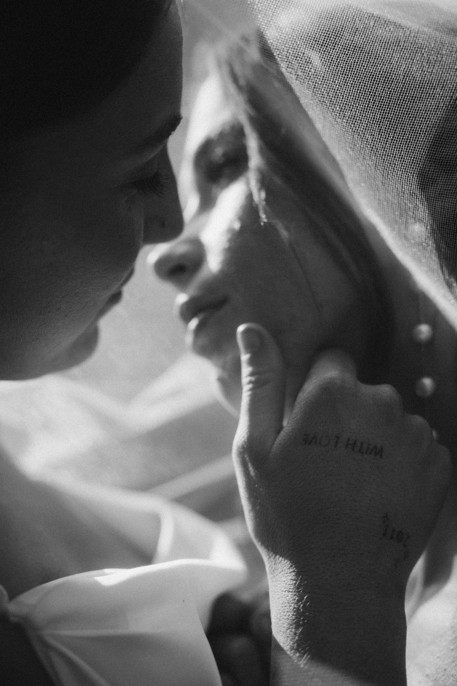 Black and white wedding photo of couple sharing intimate moment, bride in veil with groom's tattooed hand gently touching her face.