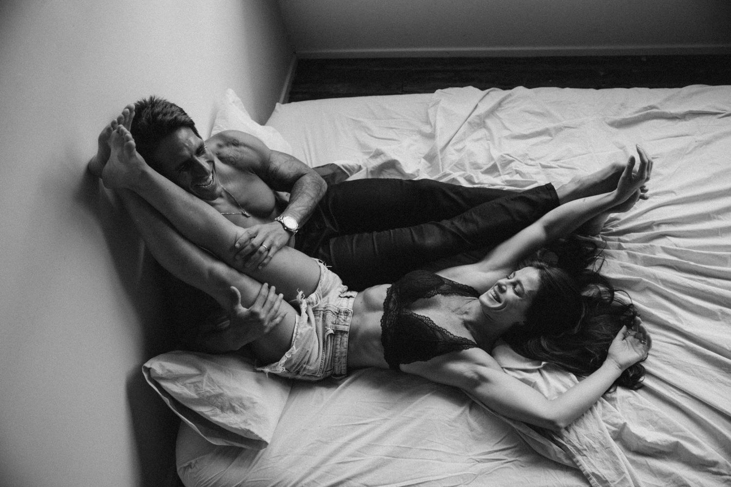 Black and white photo of a couple relaxing together on a white bed, captured in an intimate lifestyle portrait.