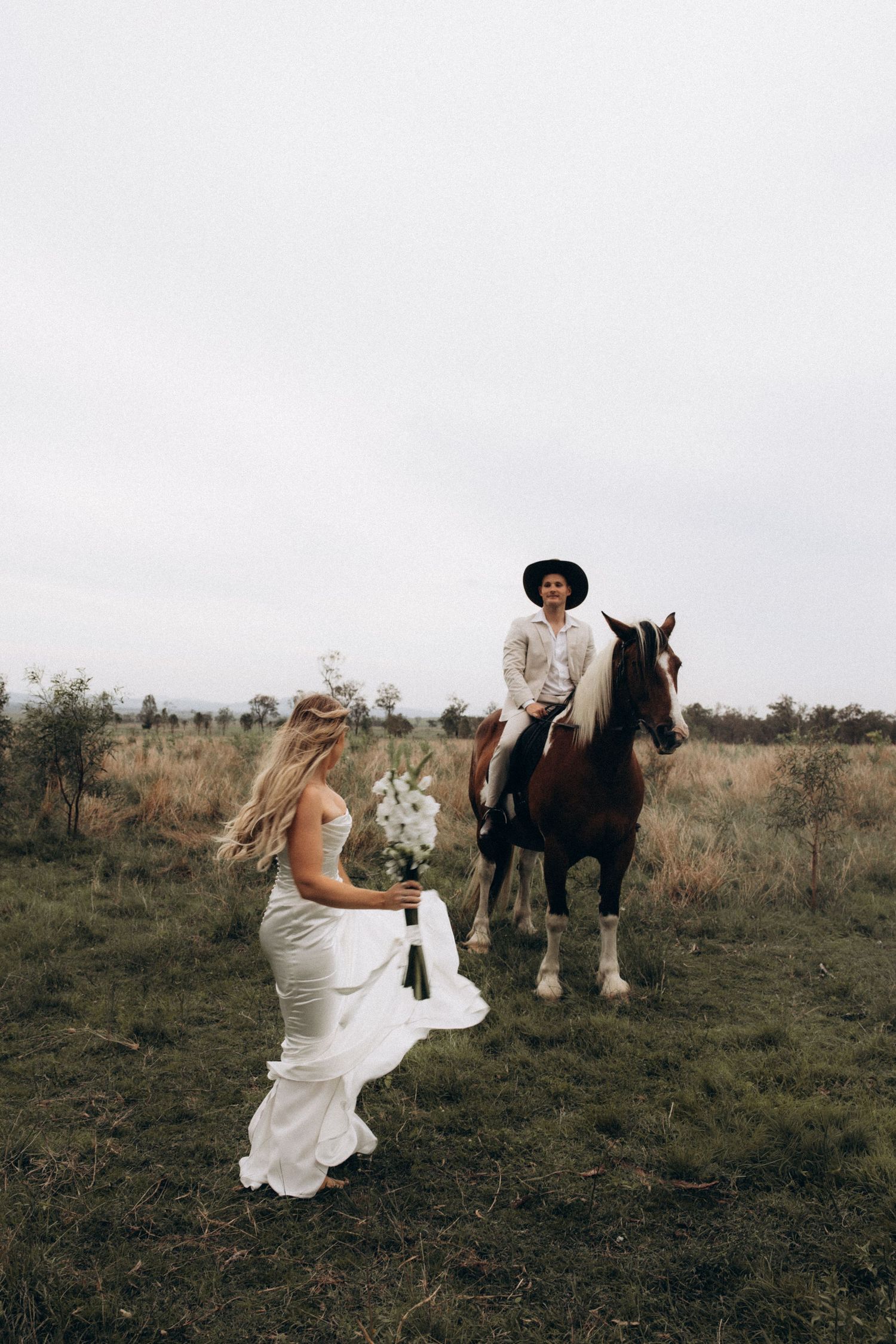 A bride in a white dress leans against a horse in a grassy field.