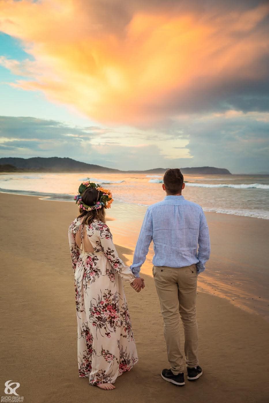 Couple holding hands on a sandy beach at sunset, bride wearing floral dress and flower crown.