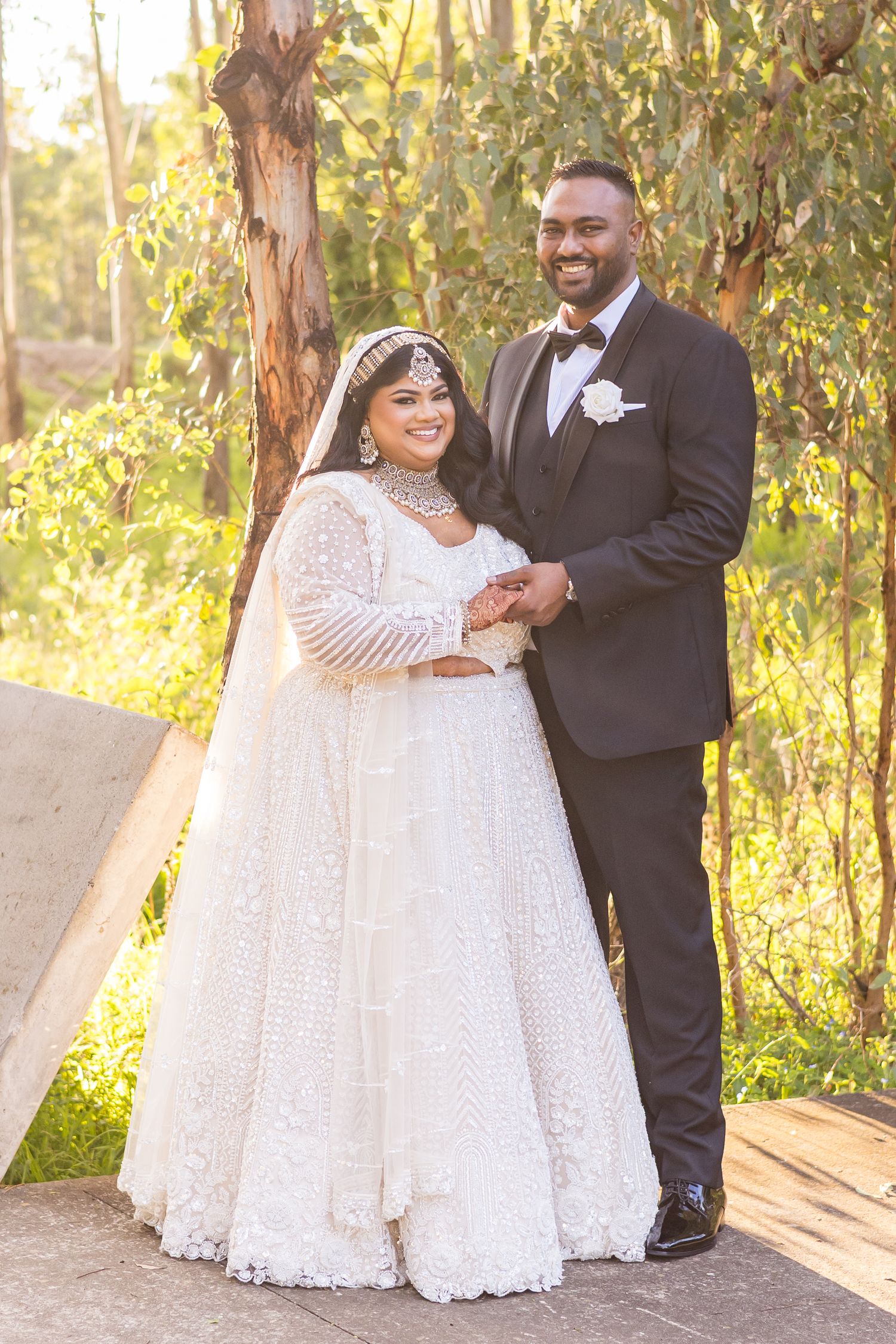 Bride in white beaded lehenga with jewelry and groom in black tuxedo posing together outdoors among trees.