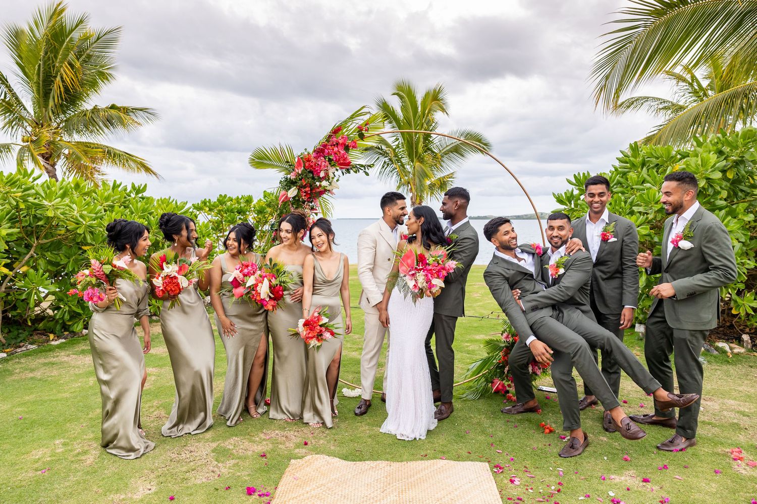 Wedding party with bridesmaids in gold dresses and groomsmen in grey suits celebrating under a floral arch outdoors.