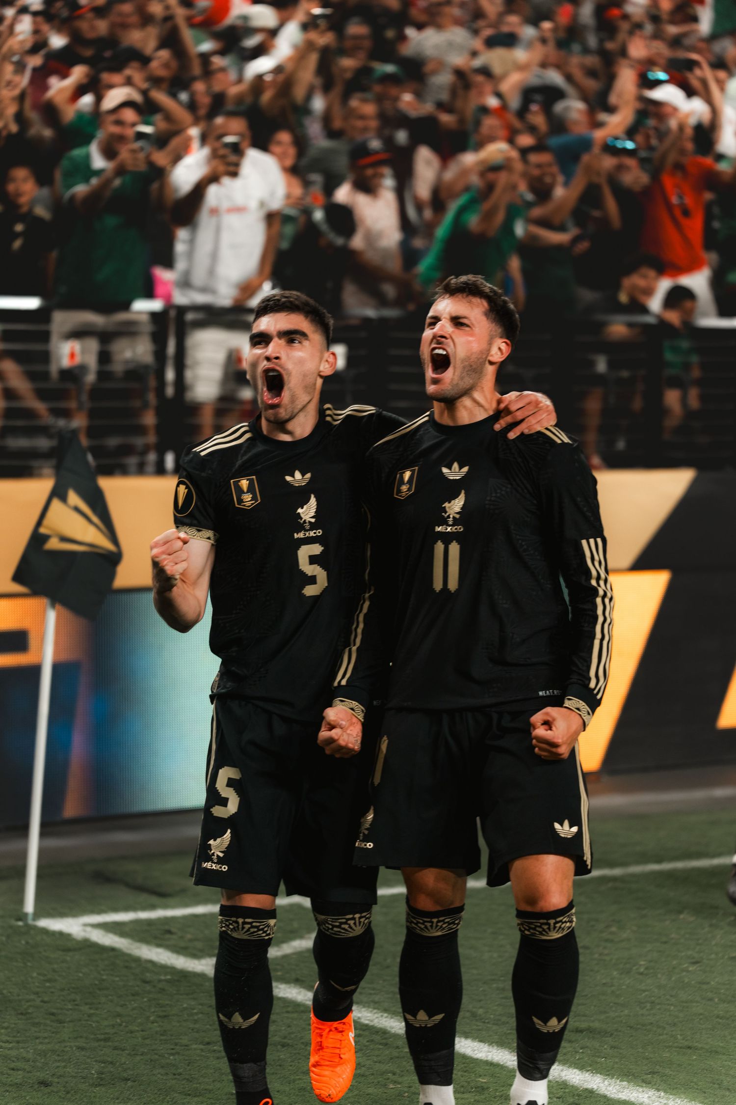Two soccer players in black uniforms celebrate passionately with fans cheering in the background during a match.