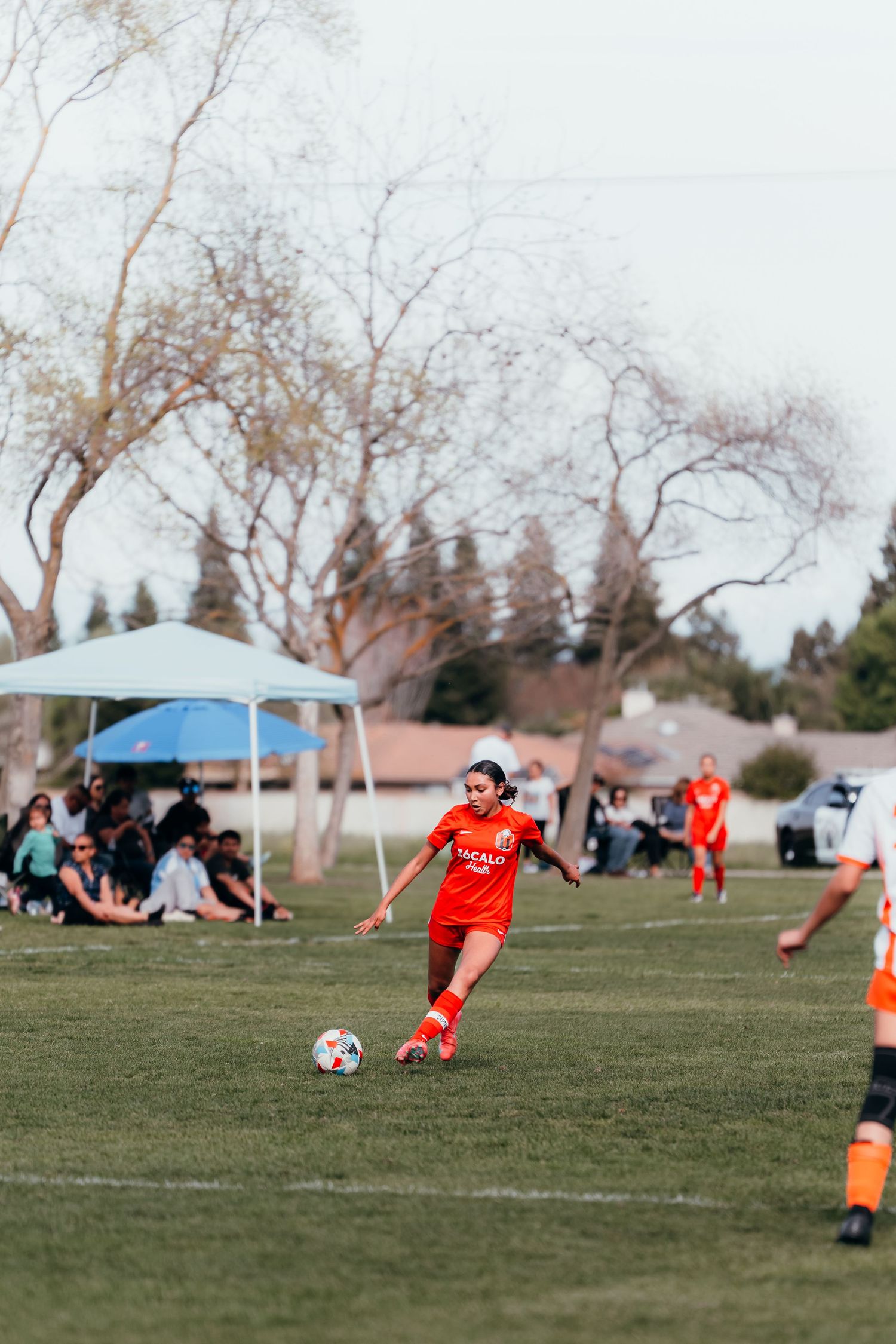 Soccer player in red jersey kicks ball on field during match with spectators under tent in background.