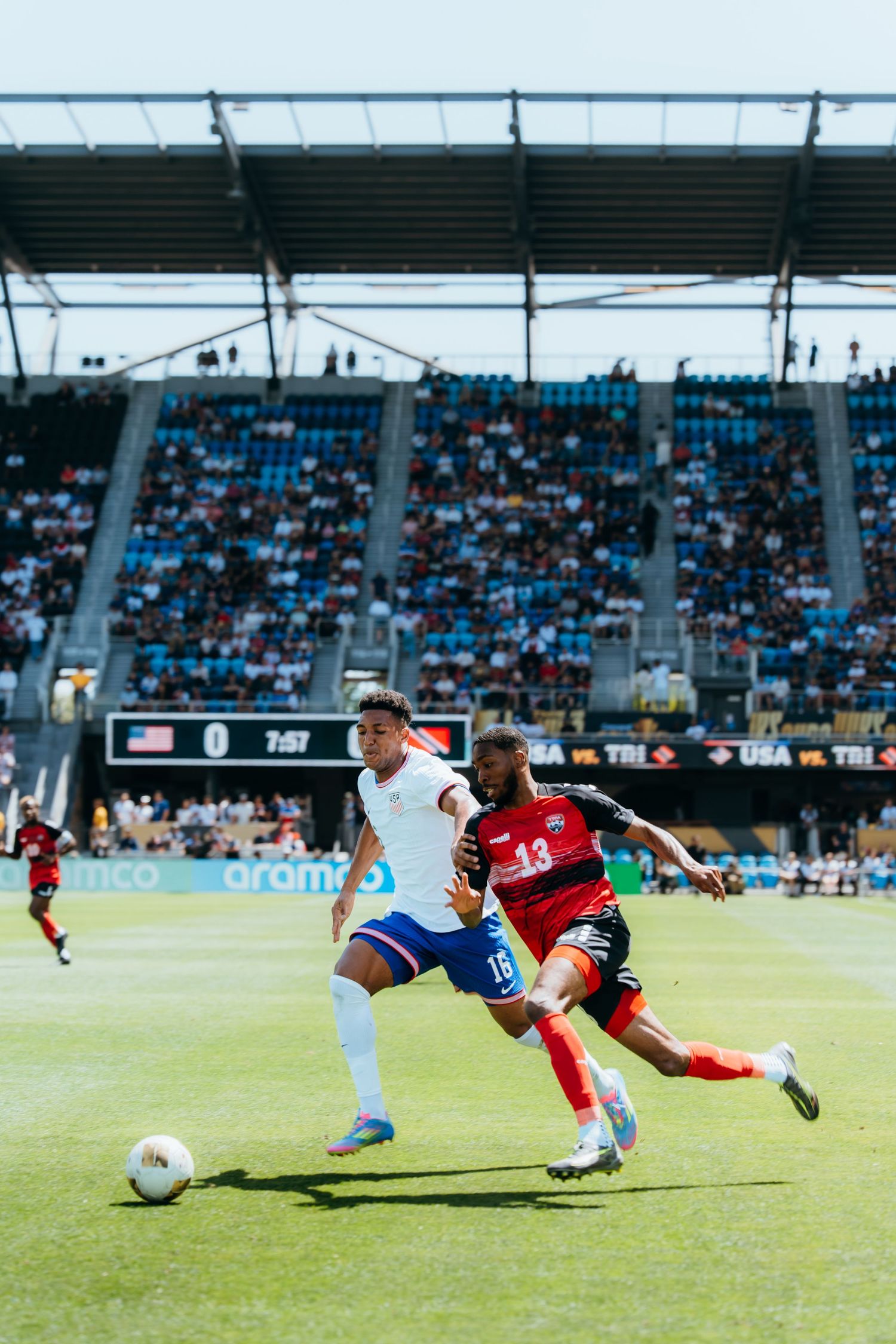 Soccer players from opposing teams compete for ball possession during match in stadium with blue seats and fans.