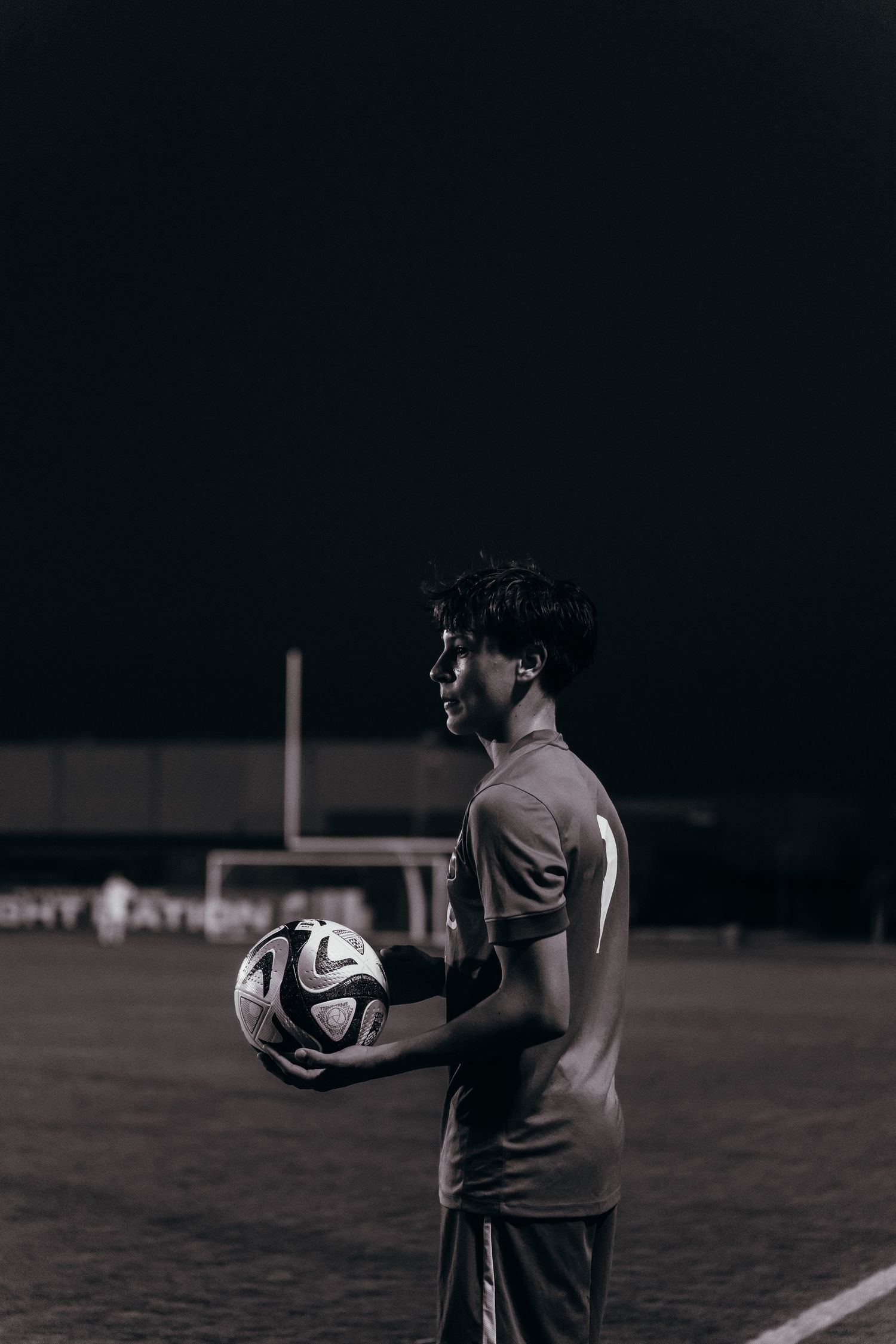 A soccer player holds a ball on a dark field at night, shown in grayscale dramatic lighting.
