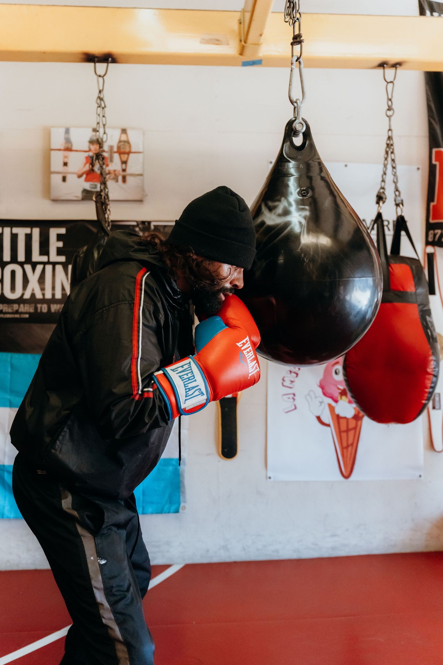 A boxer hits a black punching bag in a training gym with red gloves and black winter clothing.