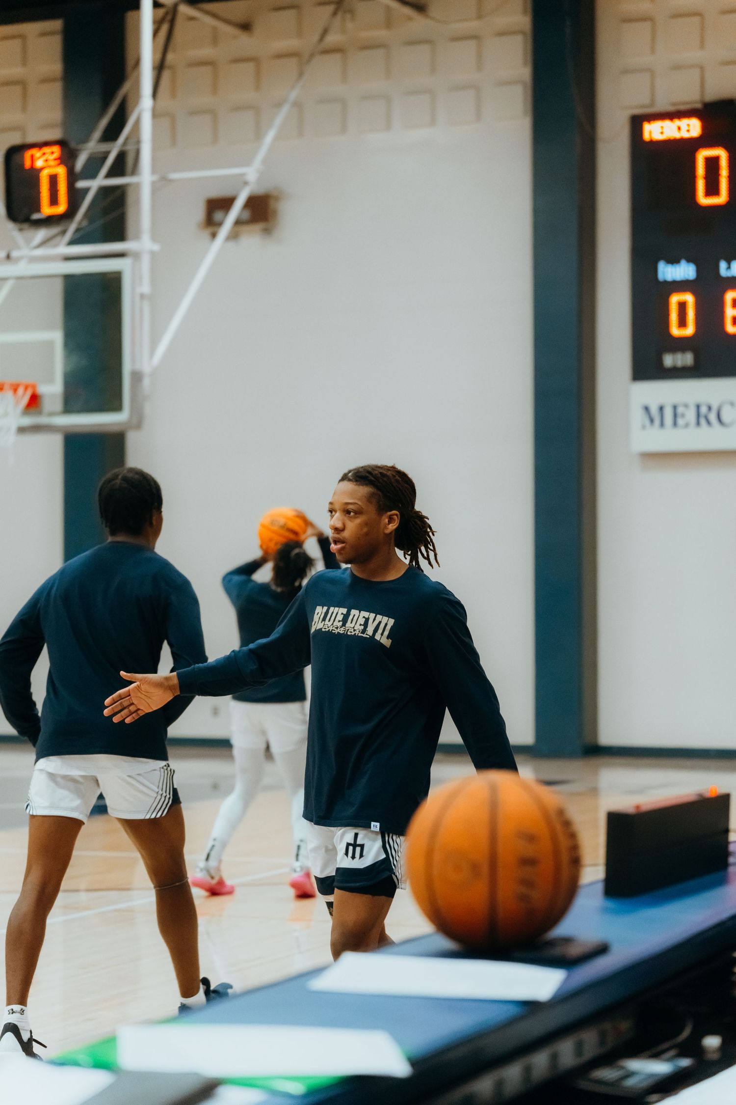 Basketball player in dark blue jersey practicing on indoor court with scoreboard visible in background.