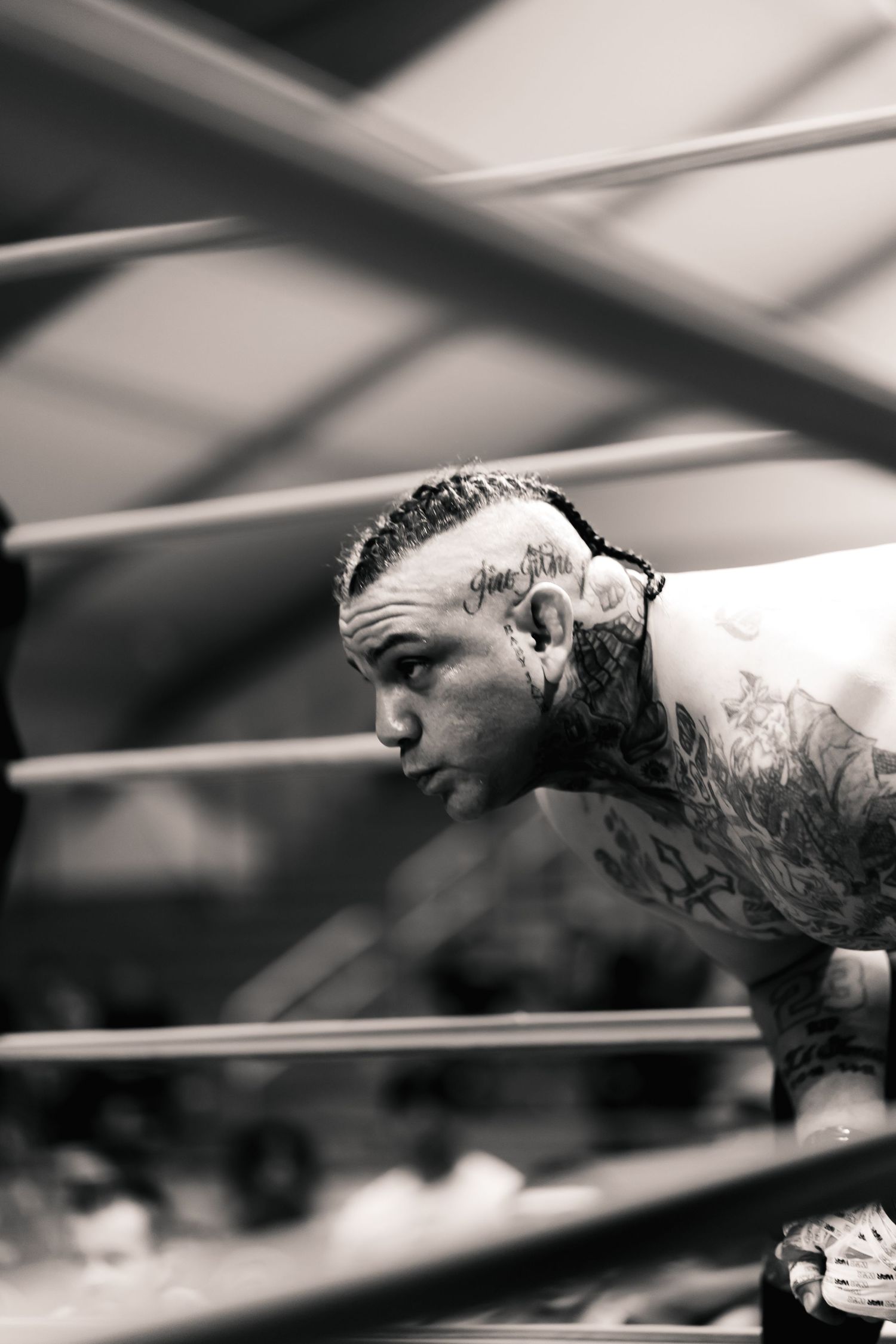 Black and white photo of a focused wrestler leaning against wrestling ring ropes during a match.