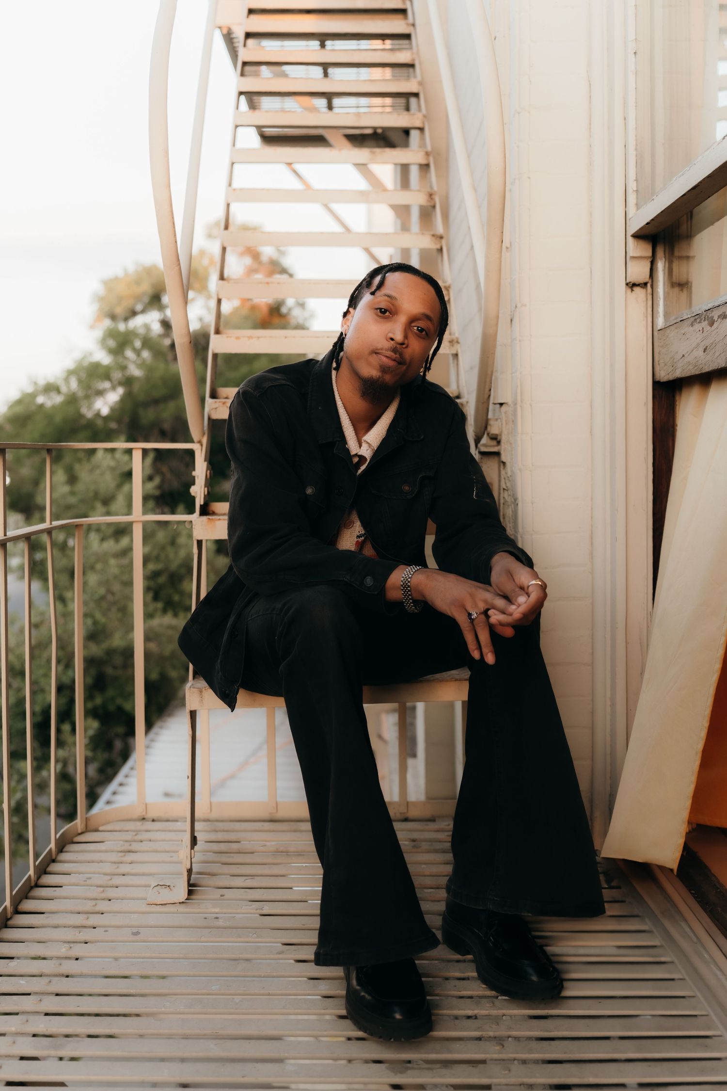 Someone sits casually on a balcony with stairs in the background wearing all black attire in warm sunset lighting.