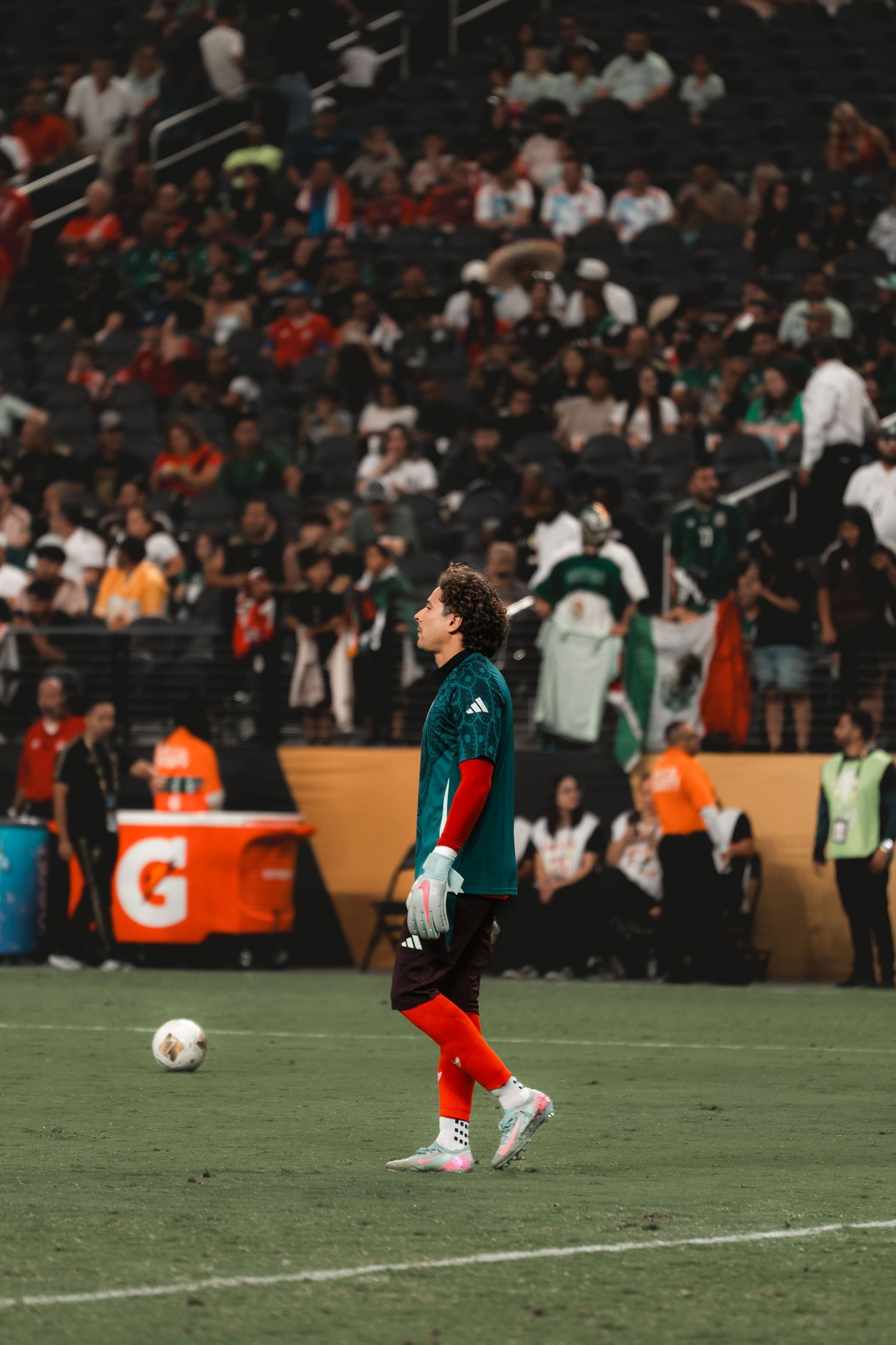 Soccer goalkeeper in teal jersey and red pants standing on field with crowd and orange coolers in background.