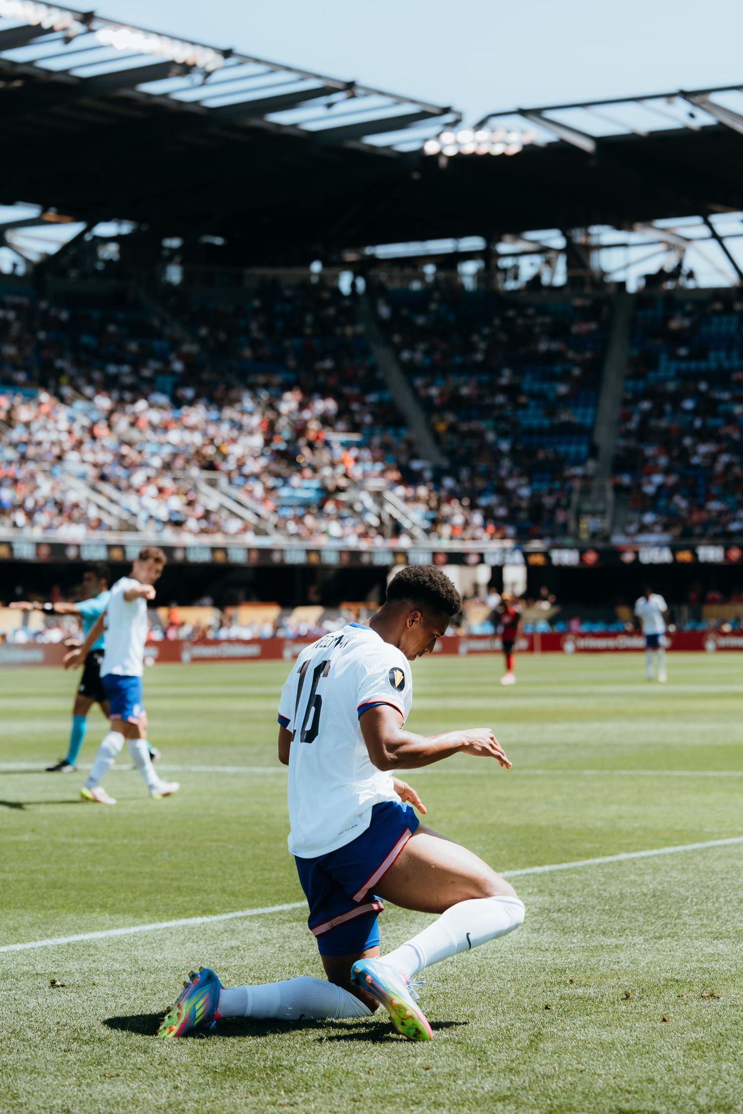 A soccer player in a white jersey slides down to the ground during a match at a crowded stadium on a sunny day.