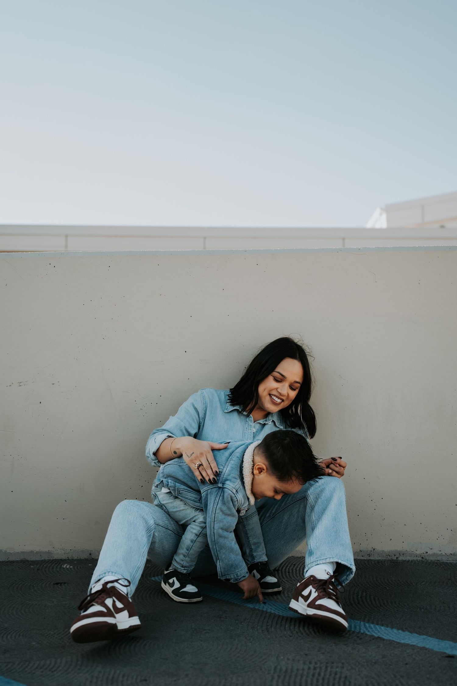 Two people in denim sitting and laughing together against white wall outdoors.