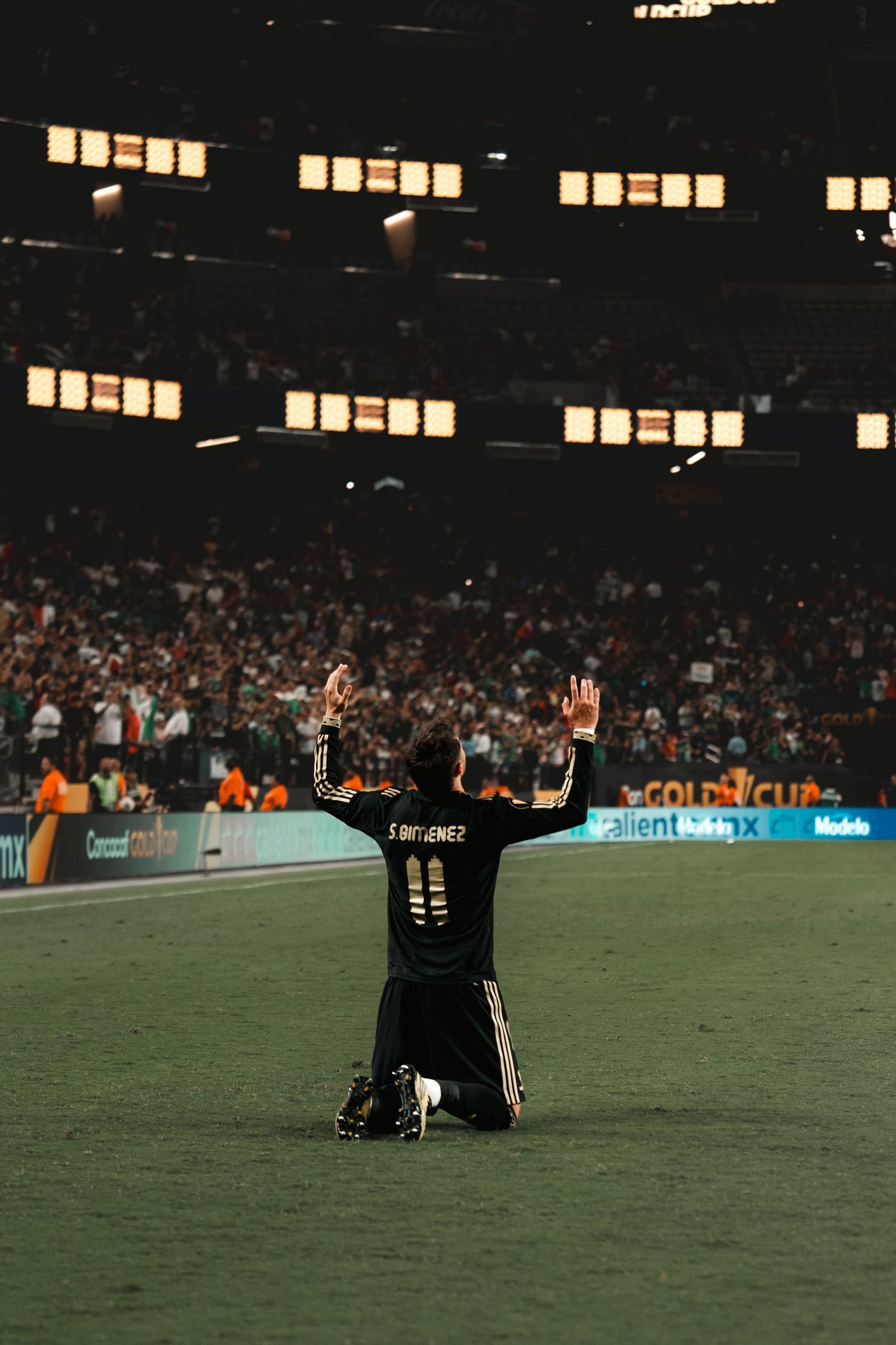 Soccer player kneels on field with arms raised in victory celebration at night stadium with illuminated windows.