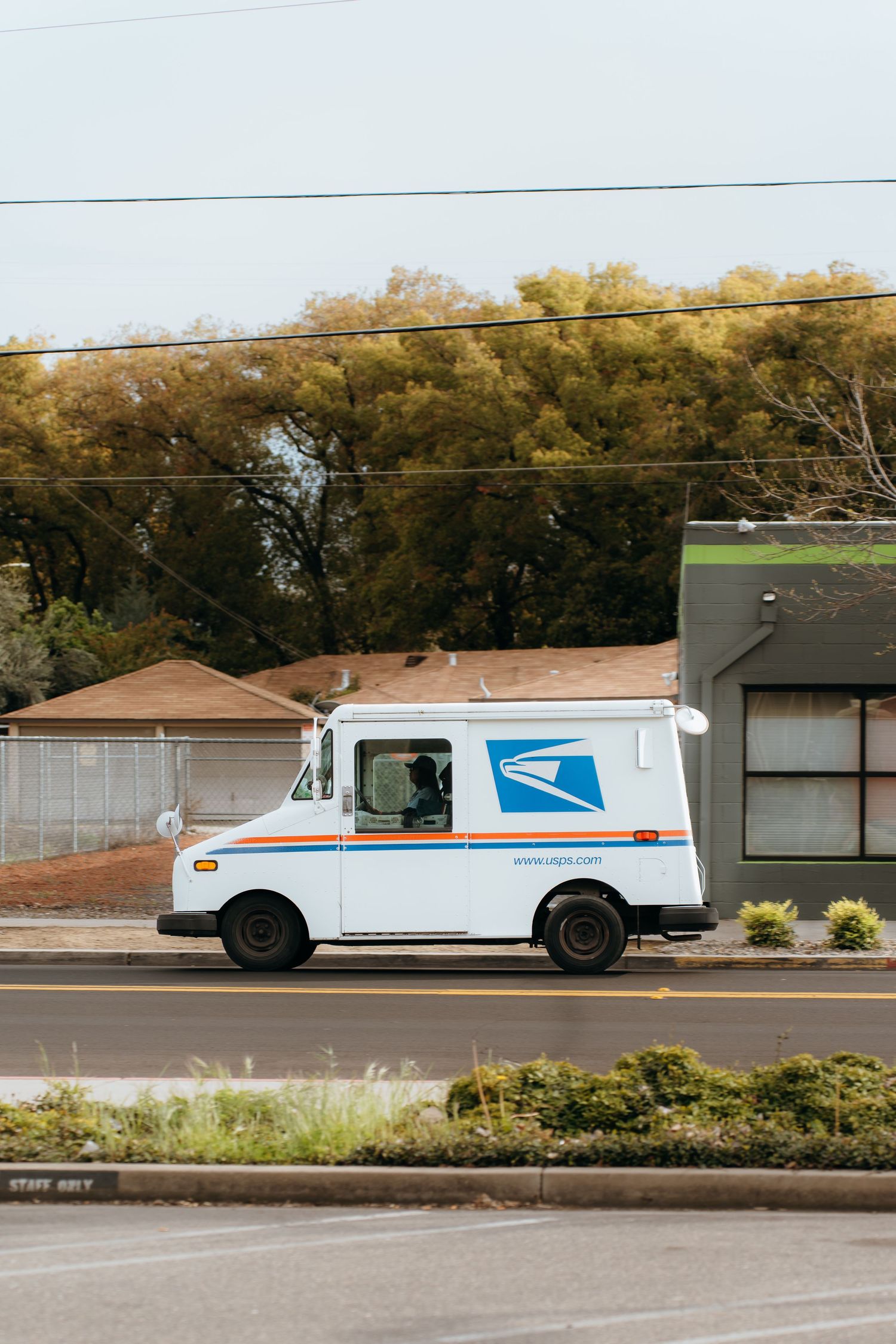 White USPS mail delivery truck parked on residential street with autumn trees in background.