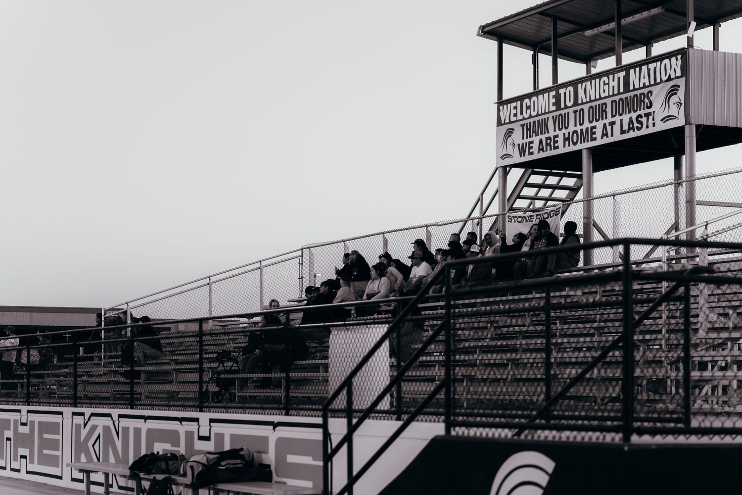 Vintage black and white photo of spectators sitting in metal bleachers at an outdoor sports venue with covered press box.