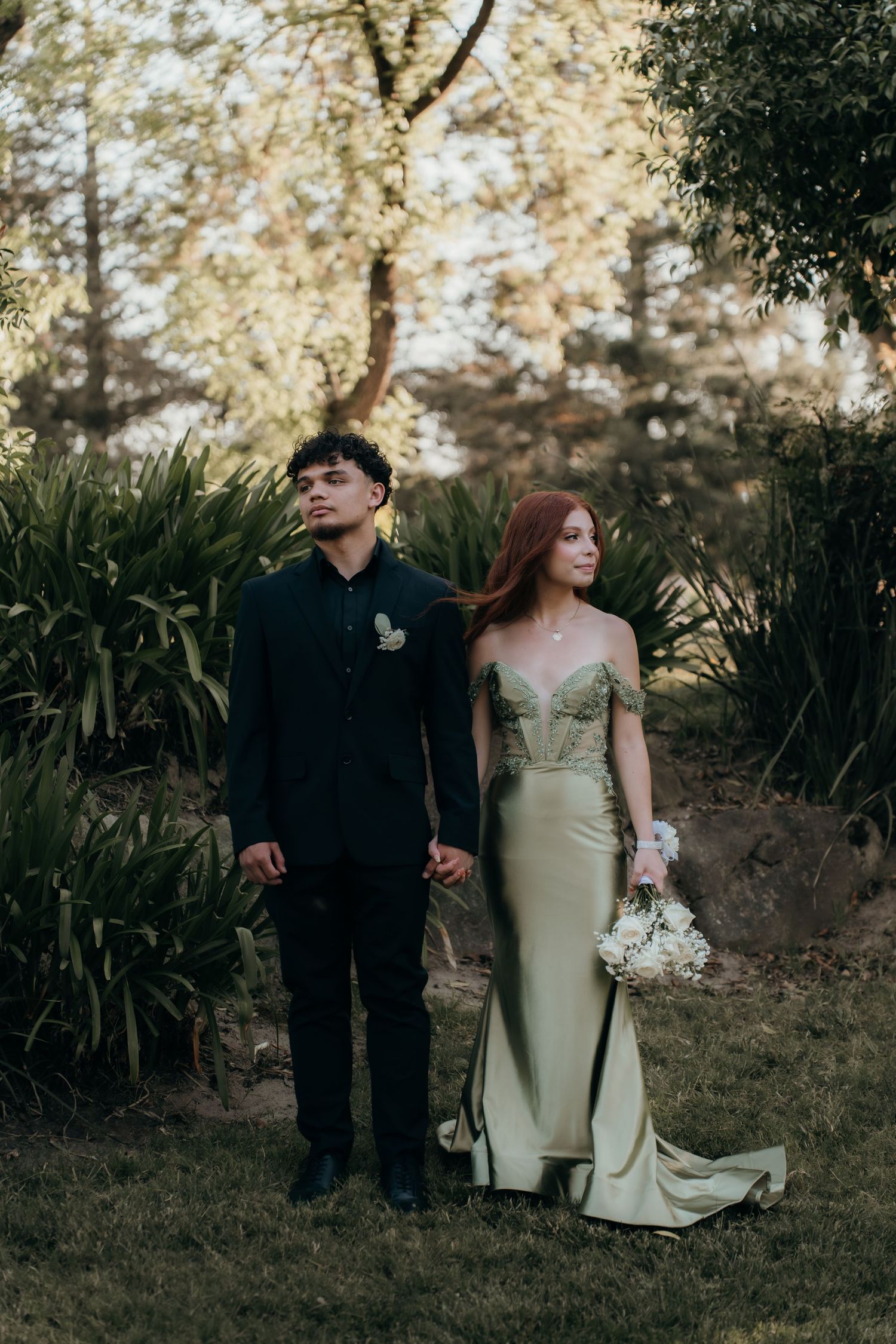 A romantic garden wedding photo with a couple in formal attire holding hands among lush greenery at sunset.
