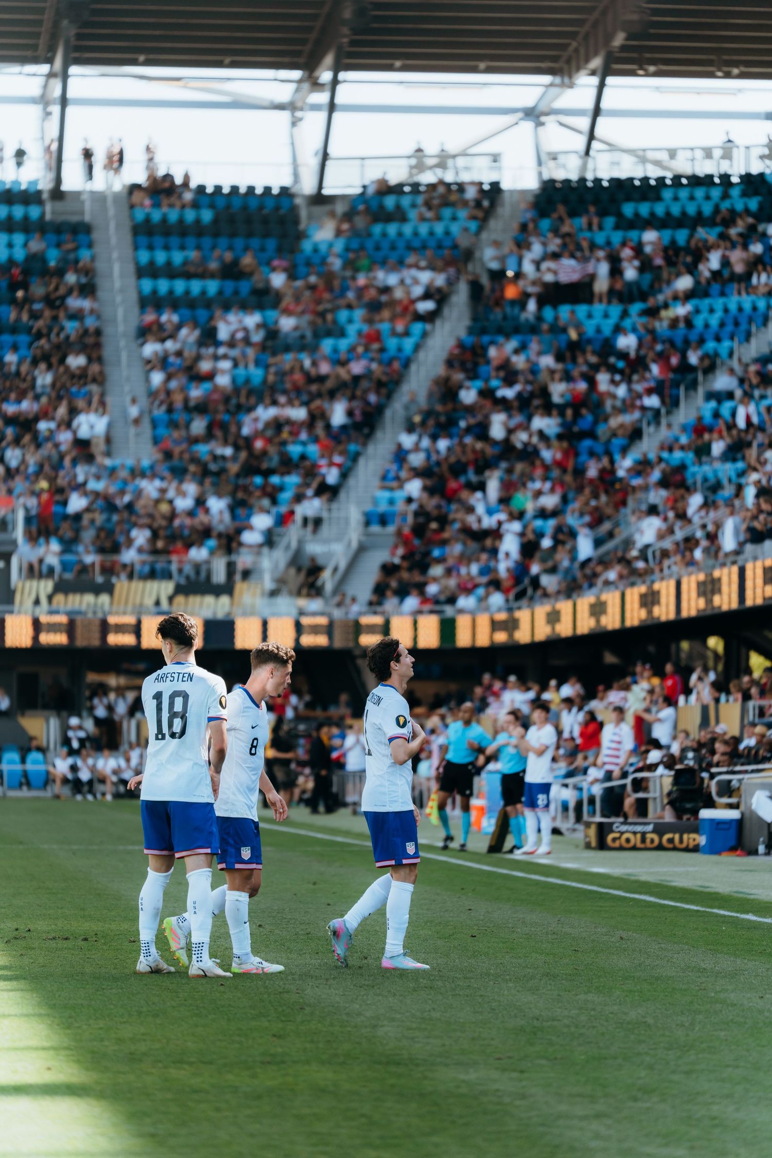 Athletes kneeling on a soccer field during a sports event with stadium seating and fans visible in the background.