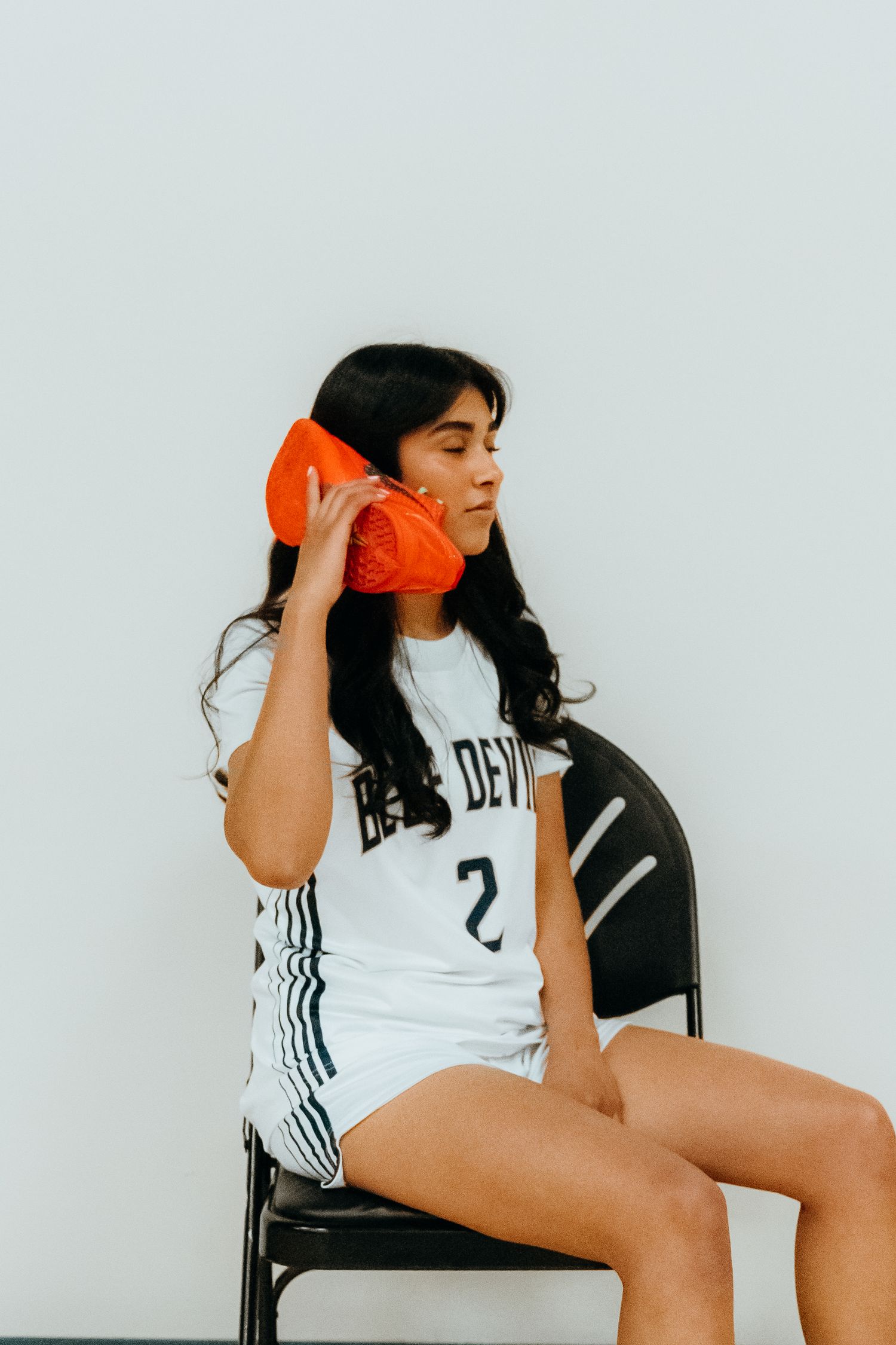 Person in white sports jersey holding orange phone to ear while seated on black chair against white wall.