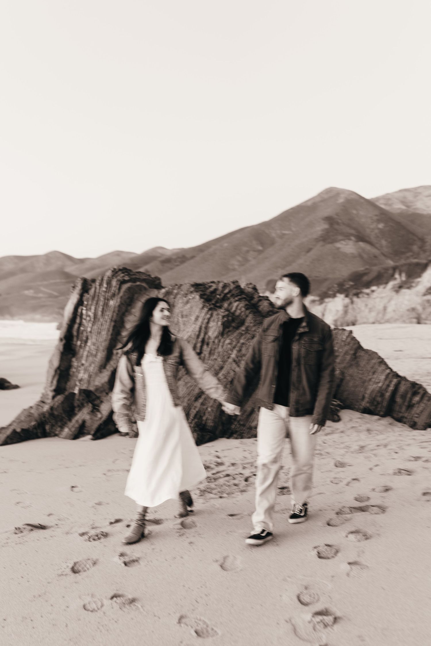 Couple walking hand in hand on beach with rocky cliffs in black and white photography.