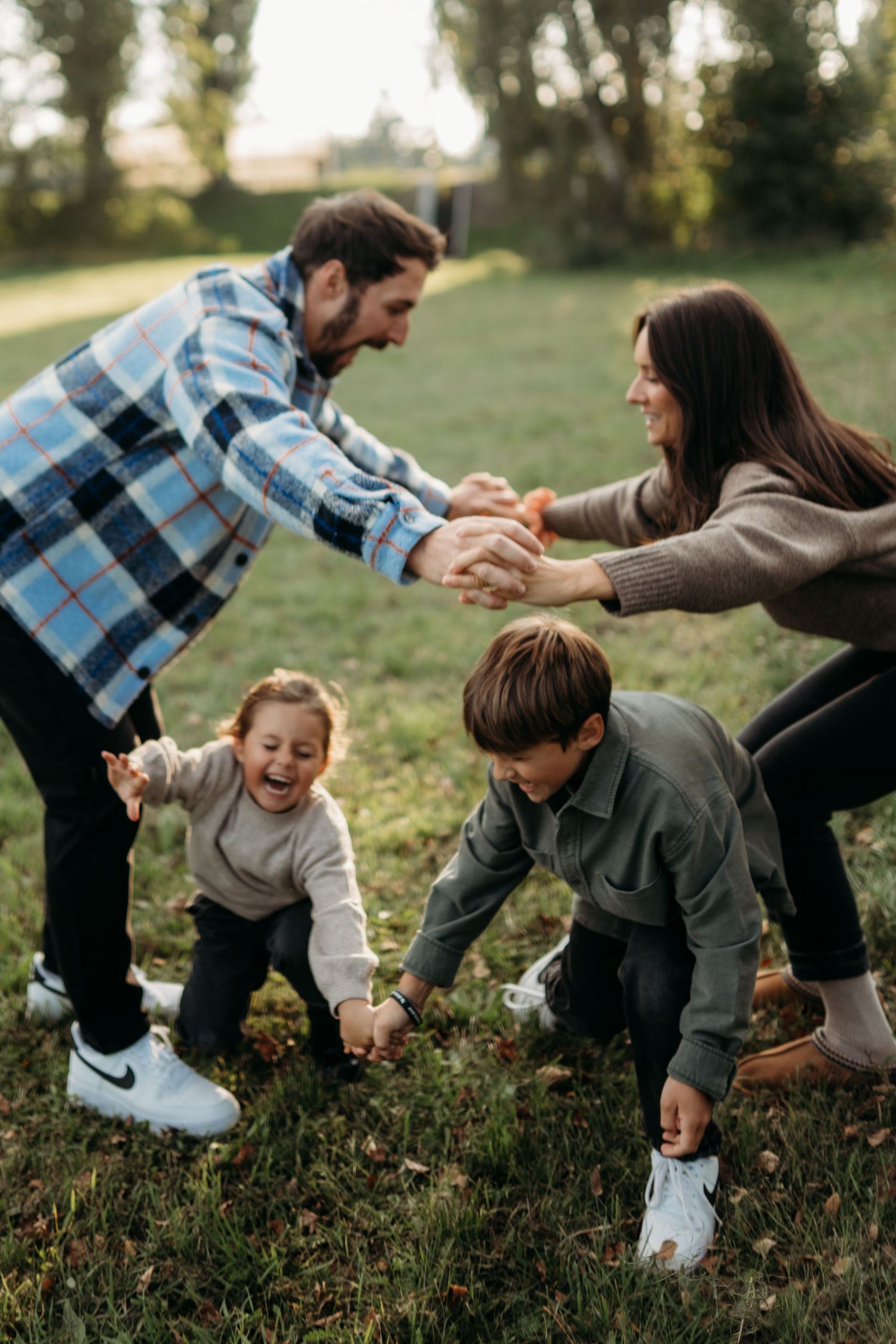 Family of four playing outdoors on green grass, parents holding hands while two young children laugh and play.
