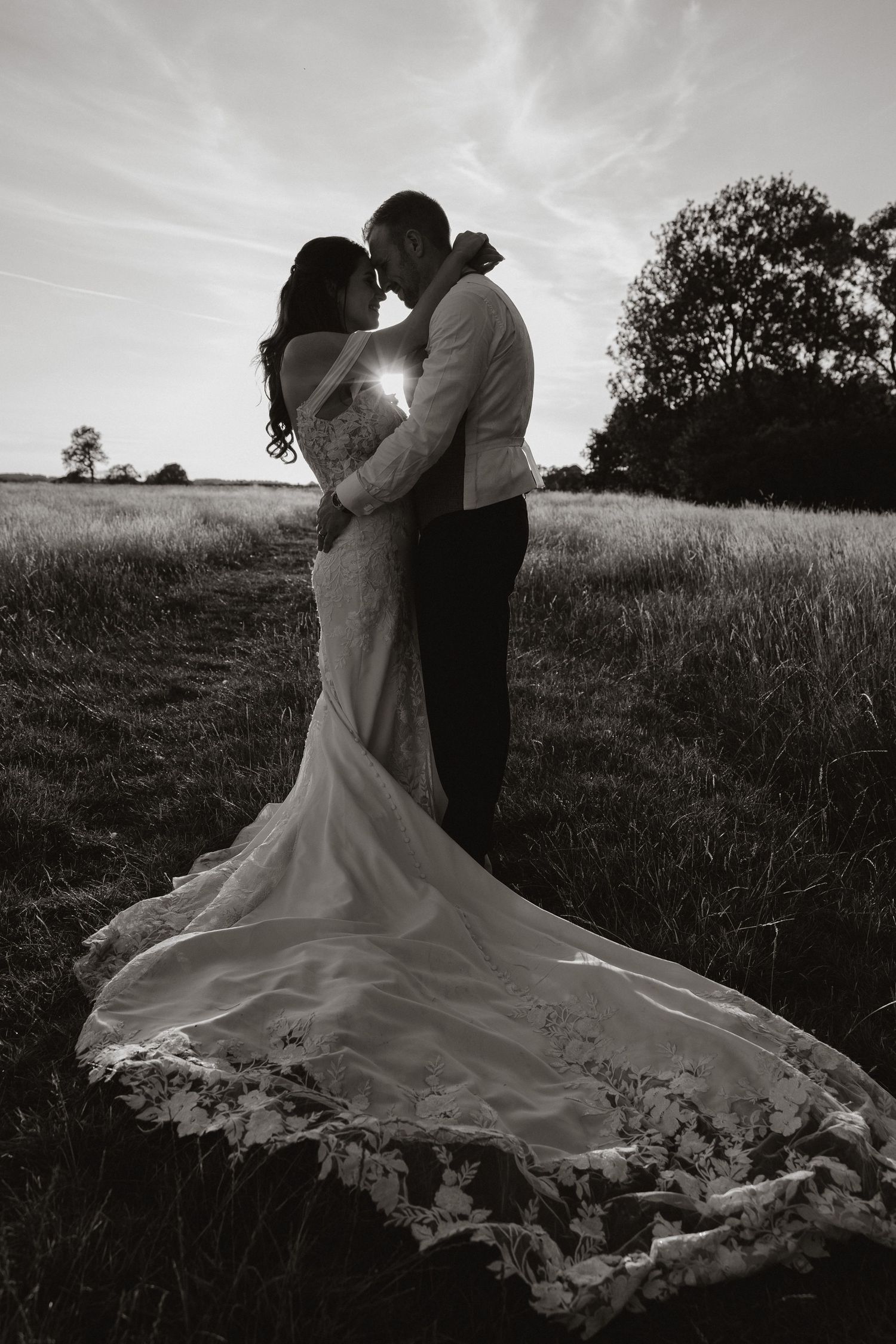 Dramatic black and white silhouette of a couple sharing a romantic kiss in a field at sunset with flowing bridal gown train.