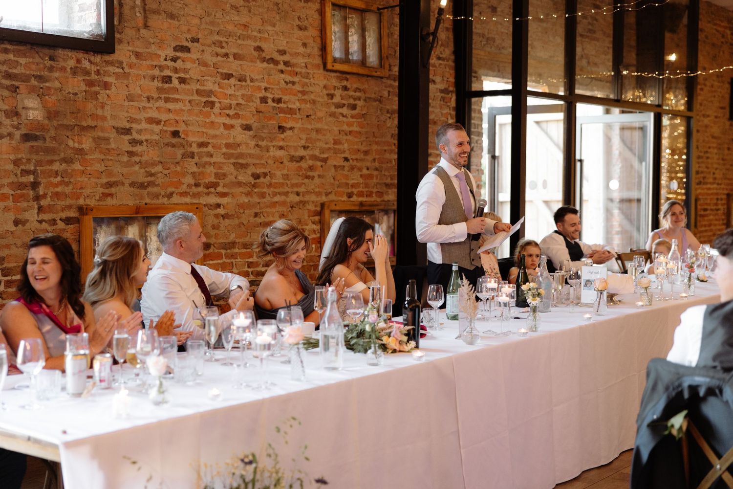 Wedding guests seated at long tables in a rustic brick-walled venue during an elegant reception celebration.