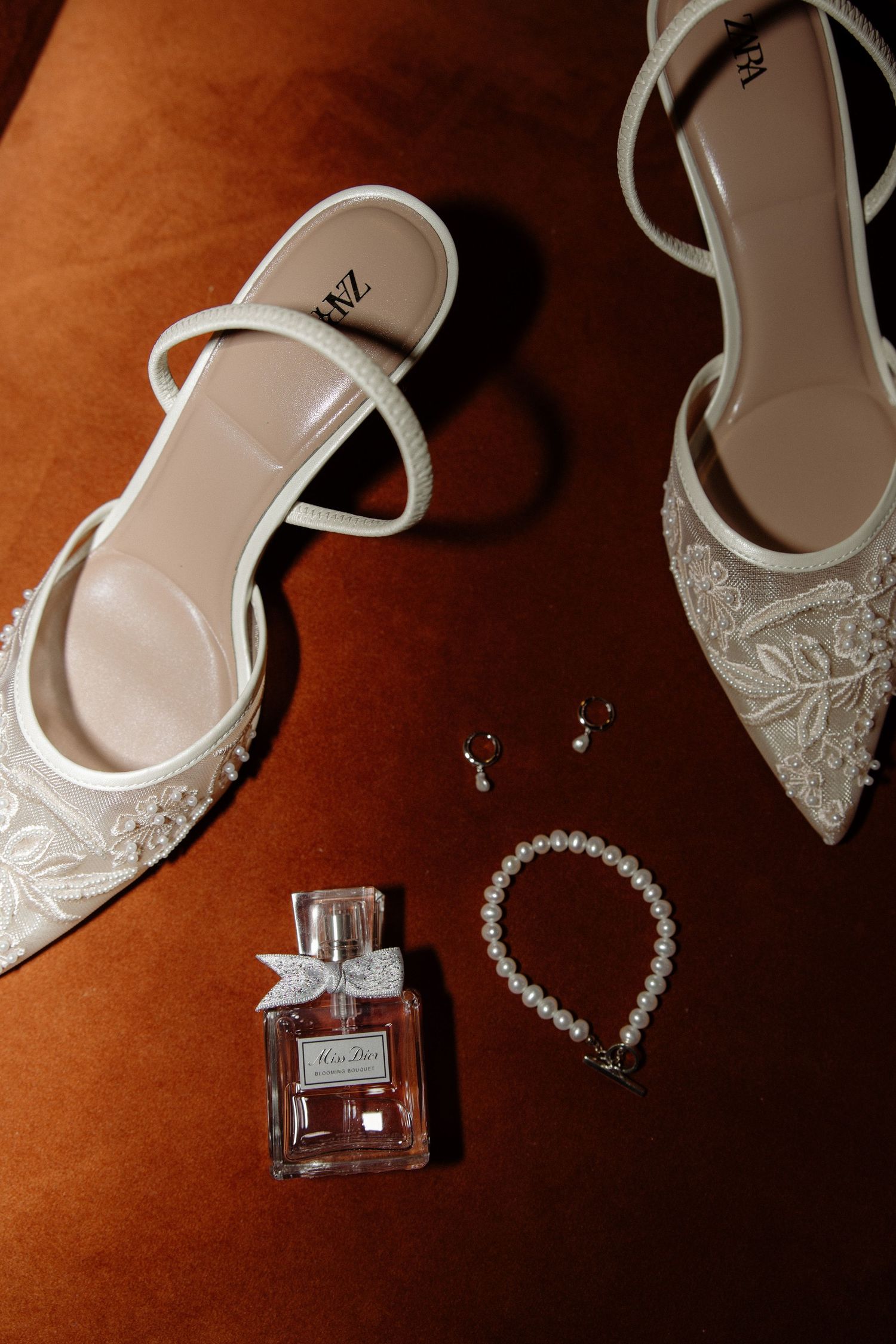 Elegant white bridal shoes with lace details displayed alongside crystal jewelry and perfume bottle on brown velvet surface.