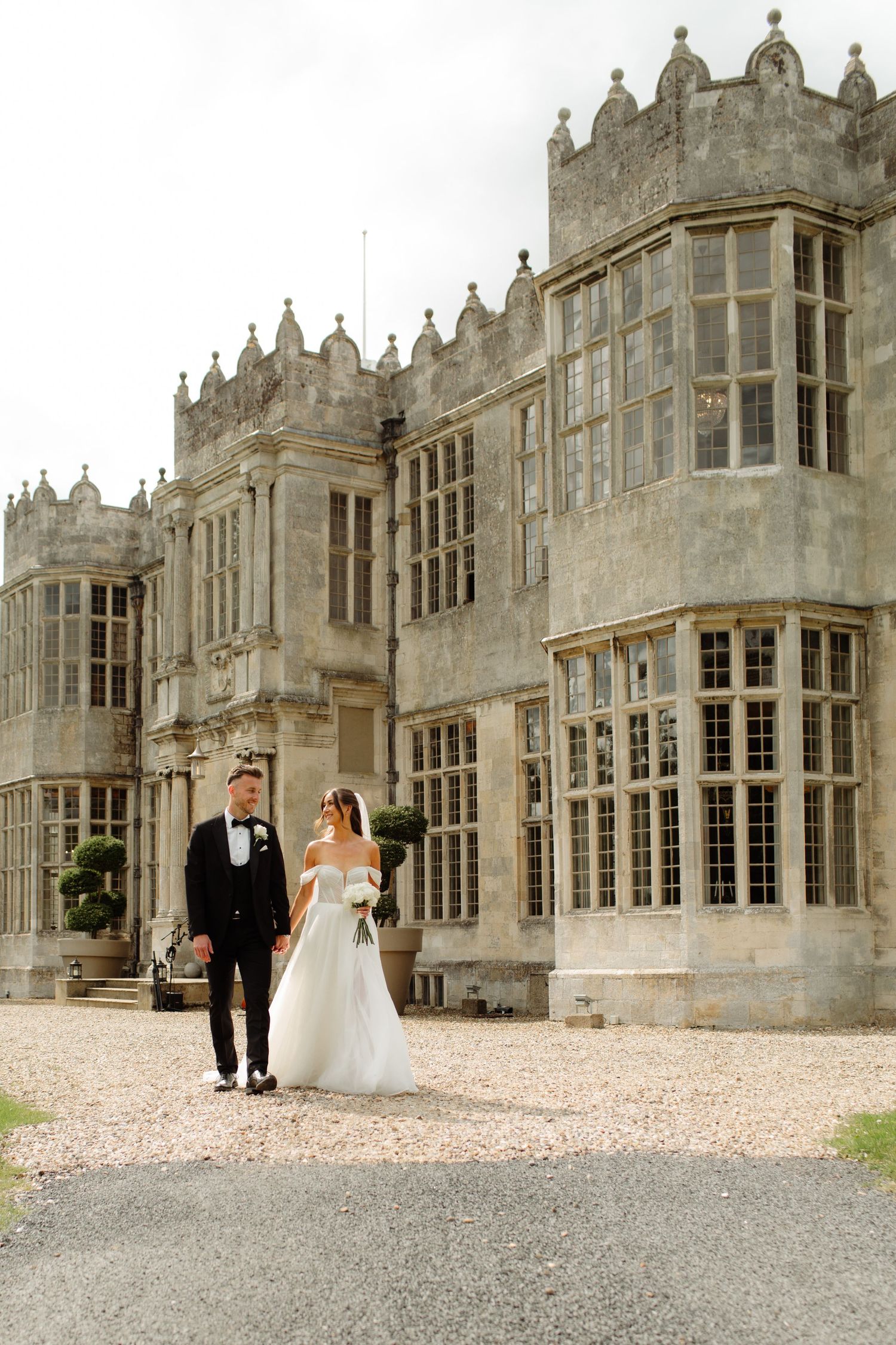 A couple walks along the gravel path outside a grand Gothic-style mansion with ornate stone architecture and bay windows.