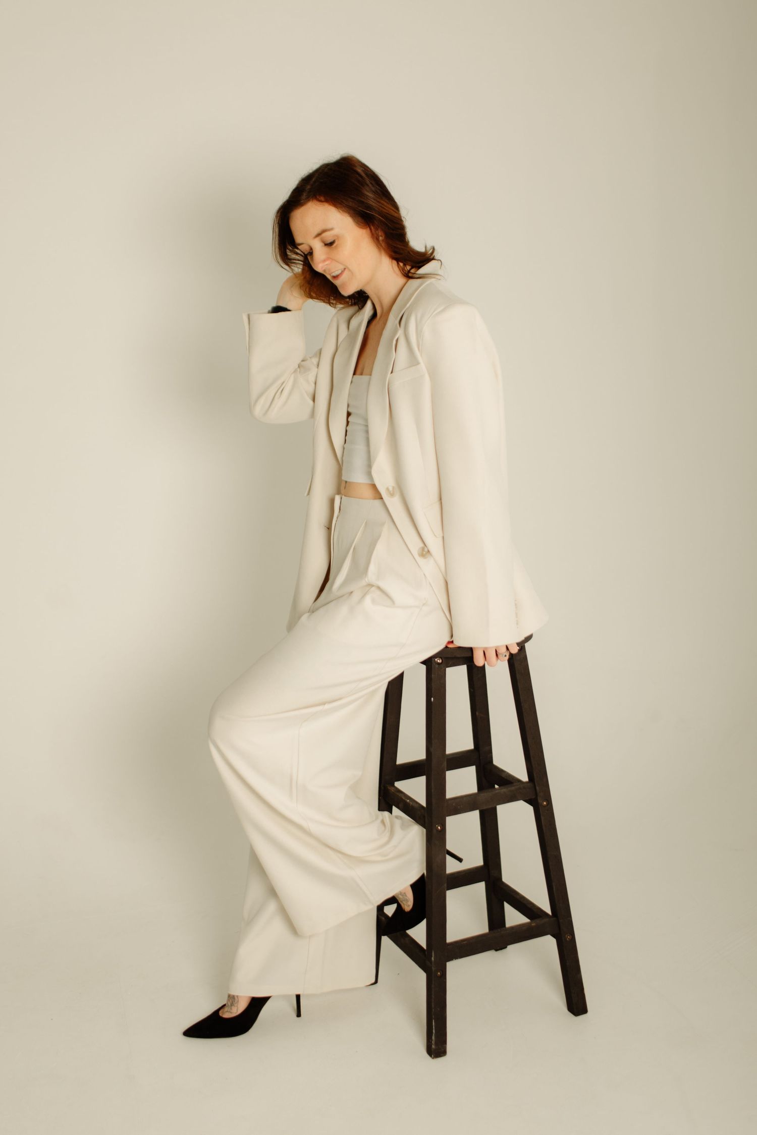 A model in a white pantsuit poses elegantly on a dark wooden stool against a neutral background.