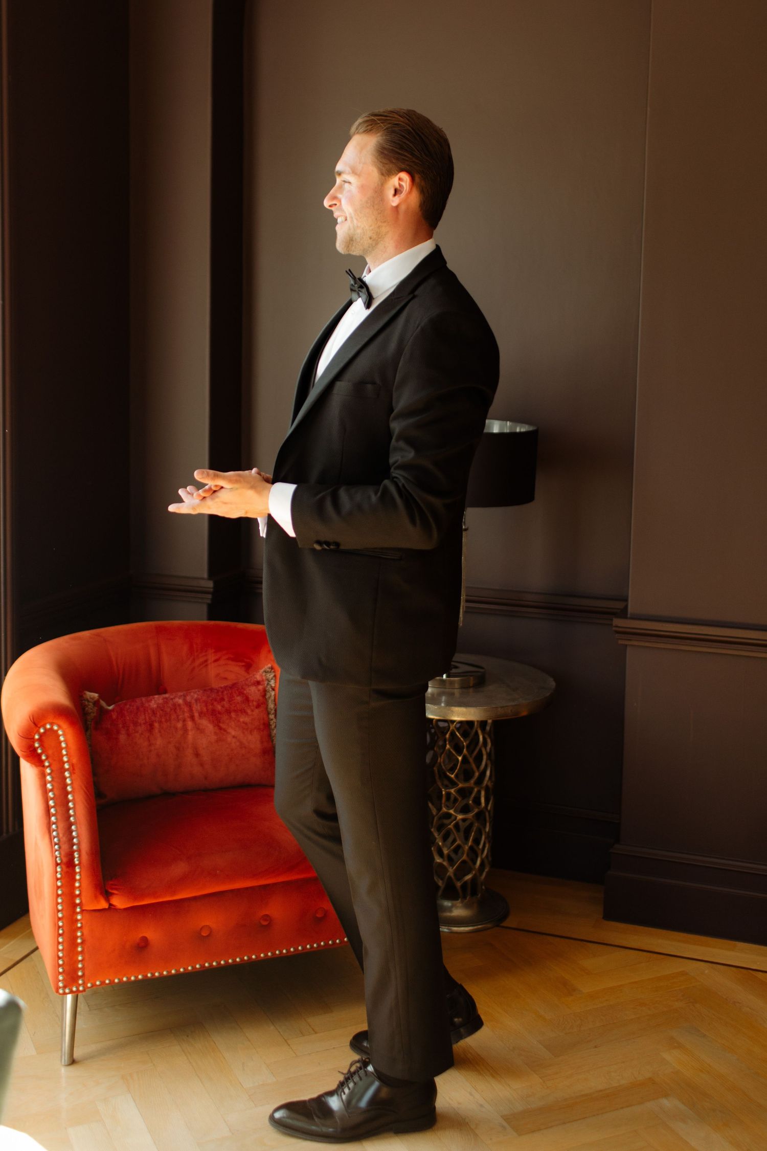 Groom in black tuxedo standing by orange velvet chair in moody lighting.