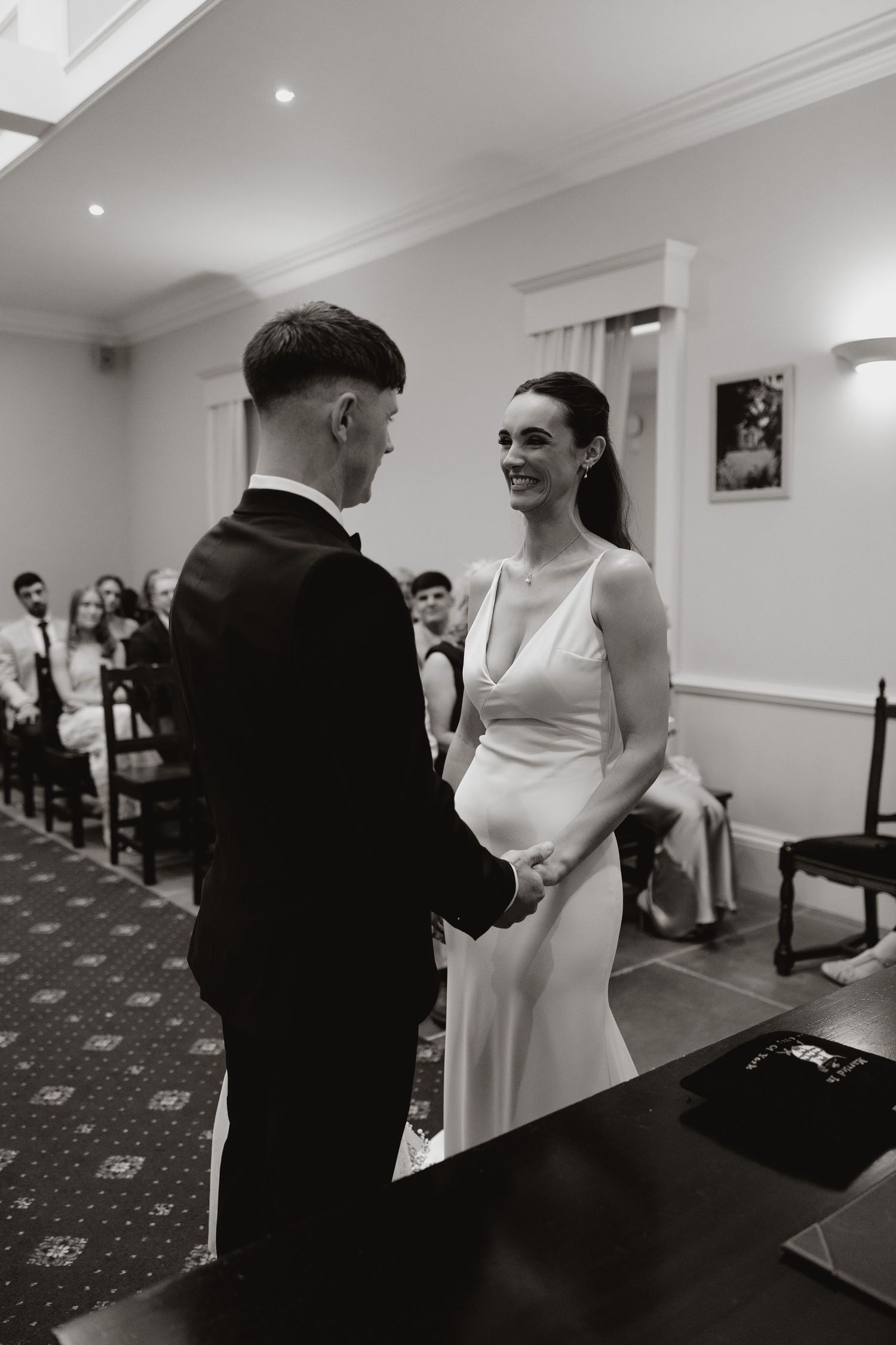A bride and groom share a joyful moment during their wedding ceremony in an elegant white room with guests looking on.