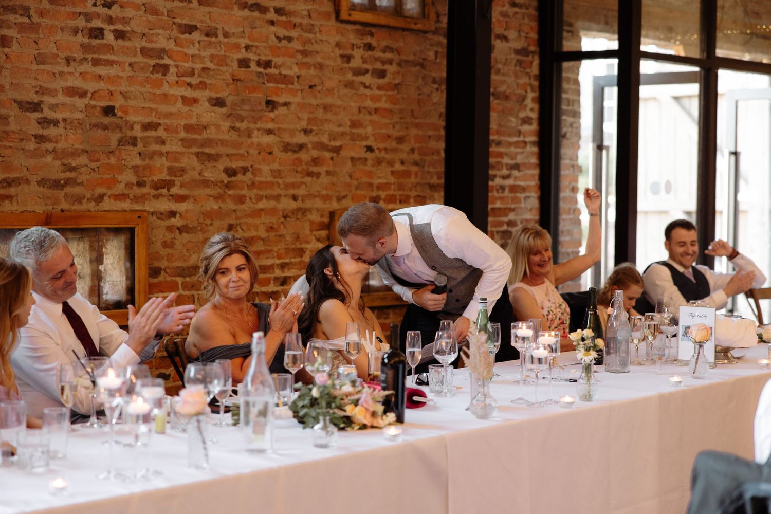 Wedding guests seated at long tables in a rustic brick-walled venue during a reception celebration.