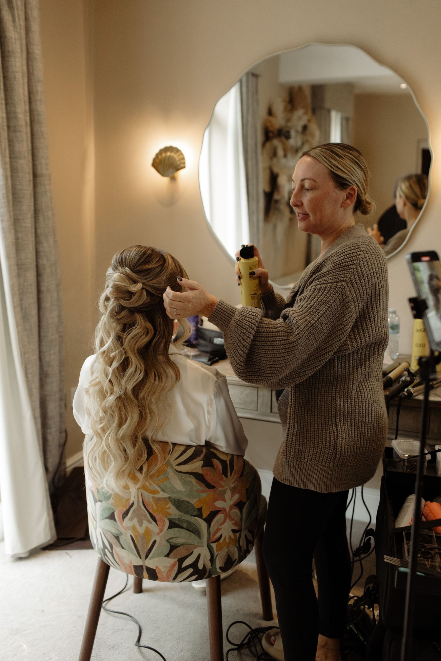 A hairstylist works on styling a bride's long wavy blonde hair in preparation for her wedding day.
