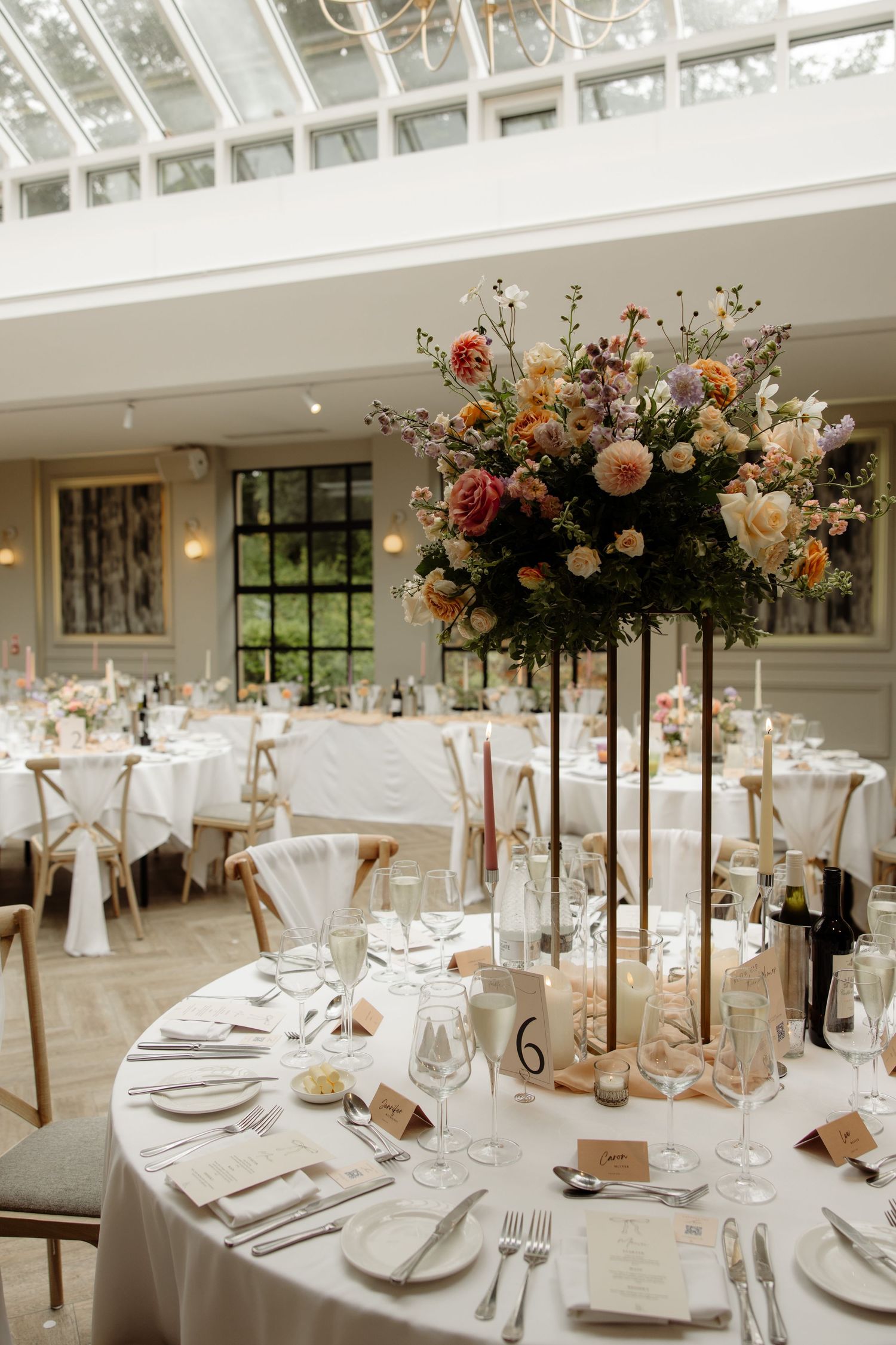 Elegant wedding reception tables decorated with tall floral centerpieces and white linens in a bright indoor venue.