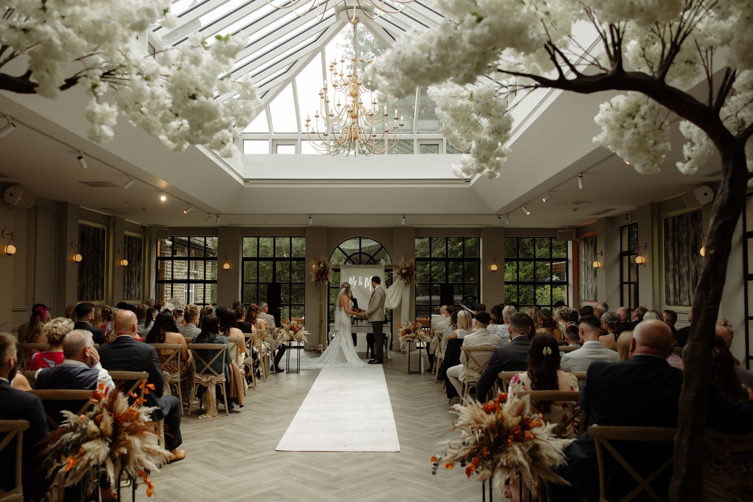 Light-filled wedding venue with glass ceilings and floral decorations creates an elegant greenhouse-style atmosphere.