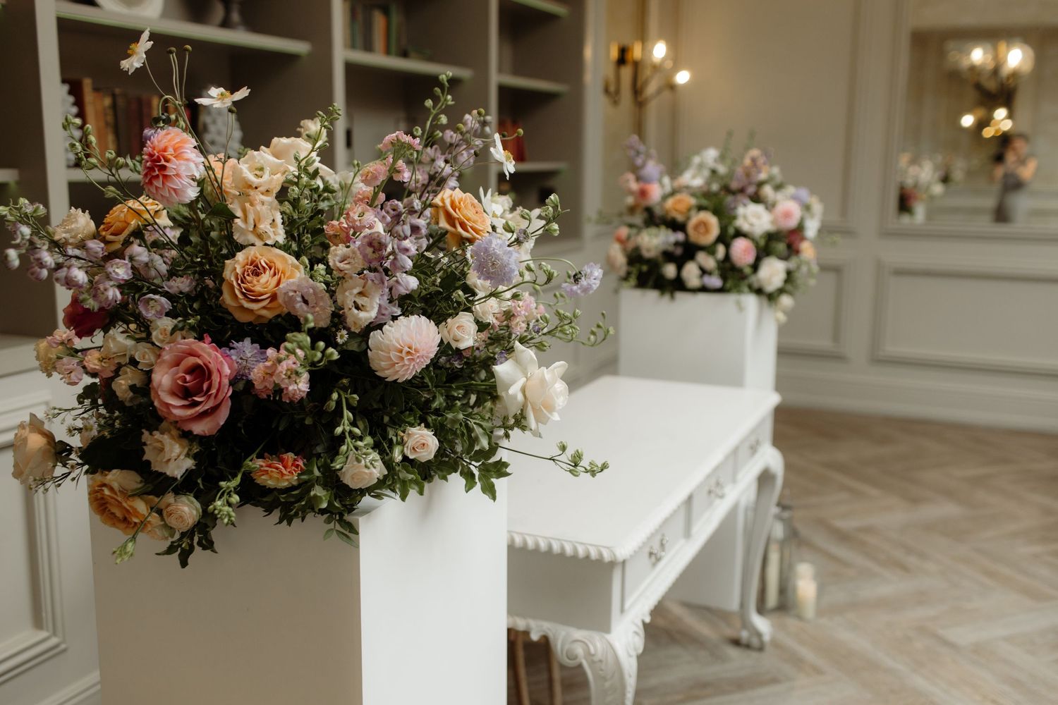 Elegant floral arrangements with peach and blush roses adorn a white wedding venue with crystal chandeliers.